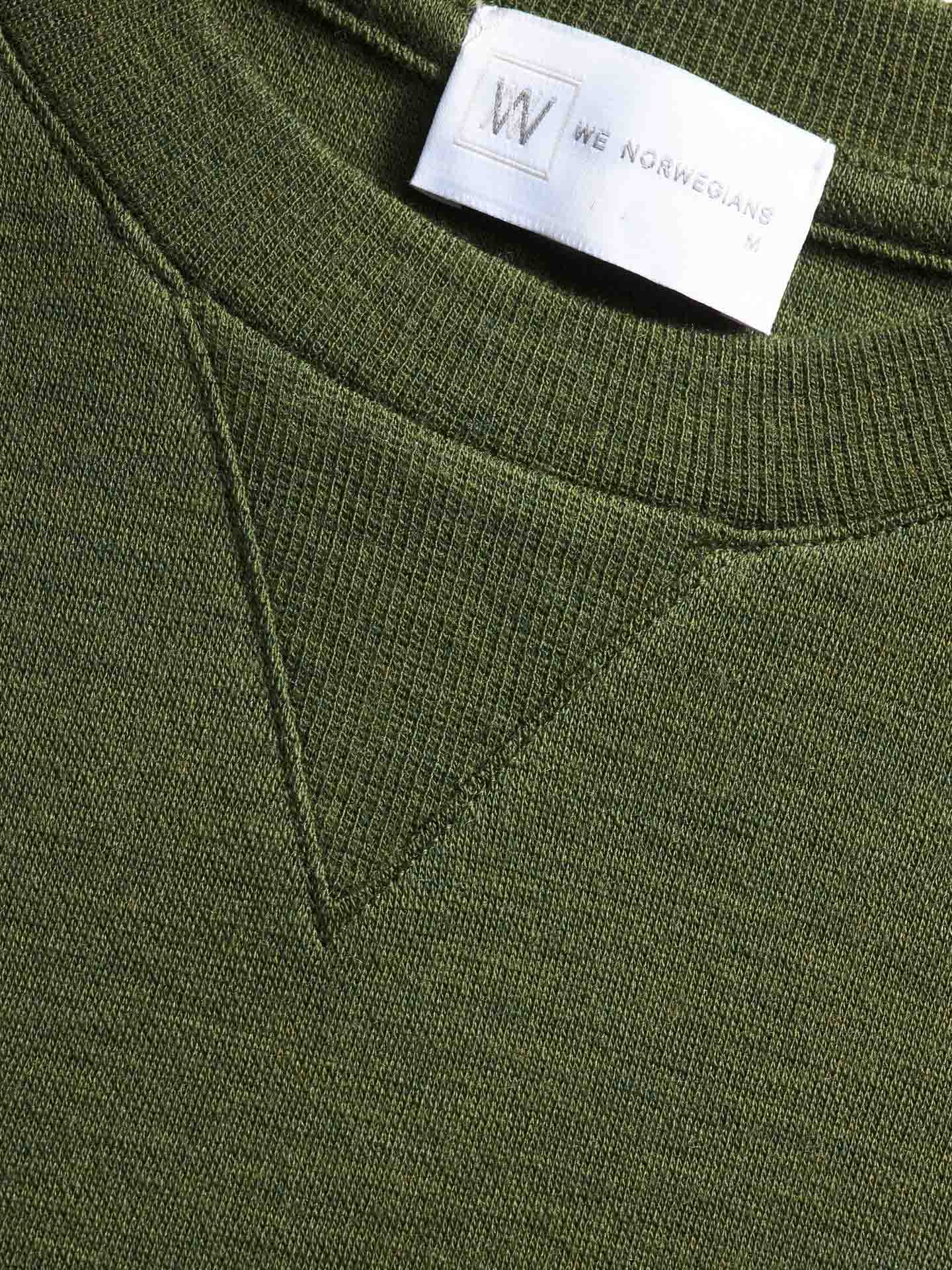 Close-up image of the olive green knitted fabric, focusing on the "Tind Crewneck Men Olive Green" sweatshirt with a triangular stitch design at the neckline. A white clothing label with the brand "We Norwegians" and size "M" is partially visible at the top, highlighting its merino wool craftsmanship.