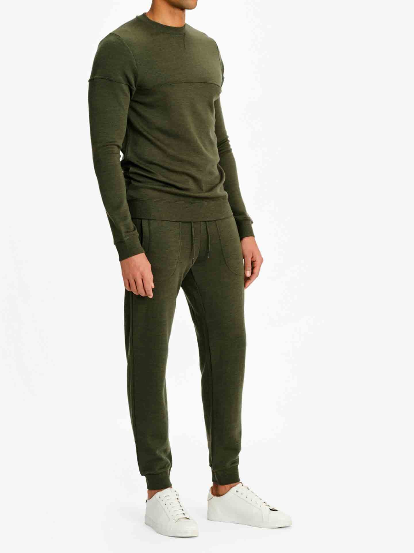 A person is wearing an olive green We Norwegians Tind Crewneck Men top and matching jogger pants with white sneakers. The outfit, reminiscent of classic sportswear, appears comfortable and casual. The background is plain white, emphasizing the clothing. The person's face is not visible.