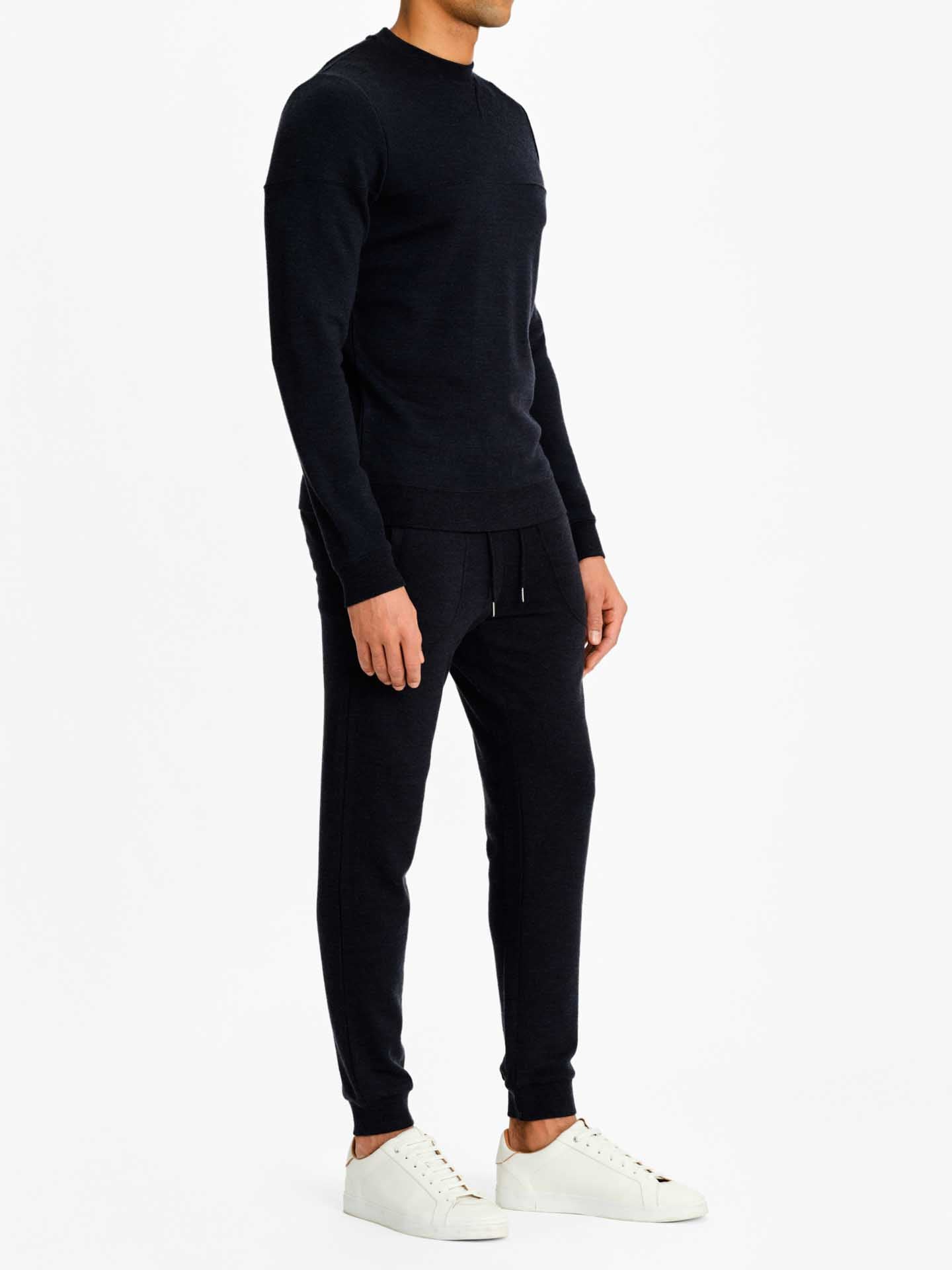 A person stands against a plain background, dressed in a matching dark-colored long-sleeve Tind Crewneck Men shirt by We Norwegians and Tind Joggers. They are also sporting white sneakers. The person's face is not visible as they are turned slightly to the side.