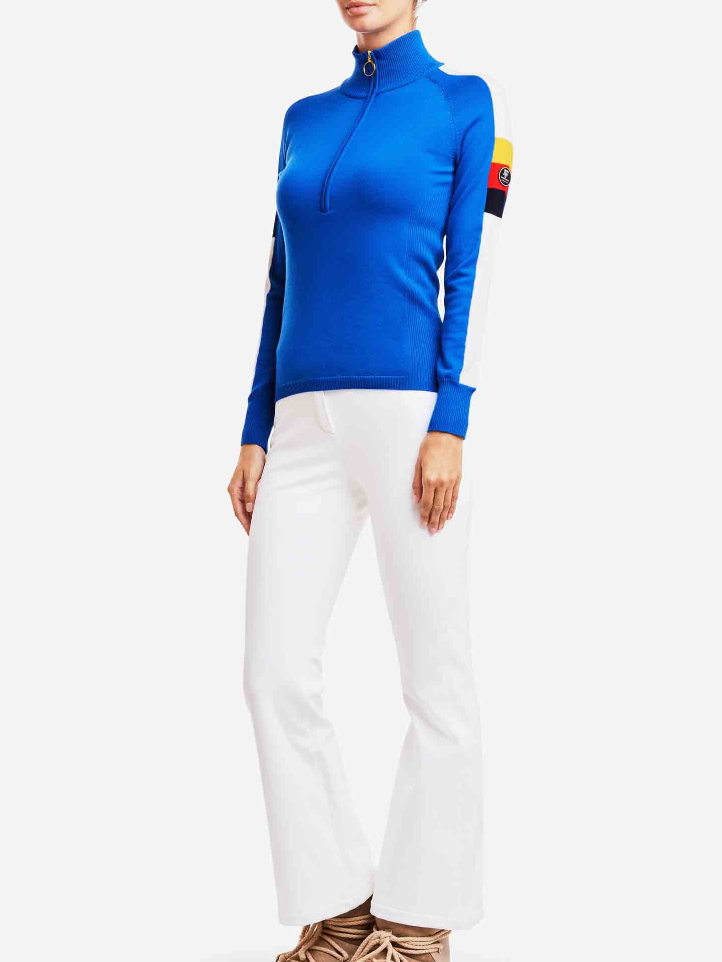 A person is wearing the Varden Zip Up Women by We Norwegians, a blue long-sleeve zip-up top with a patch on the left arm, paired with white flared pants reminiscent of 70s inspired ski fashion. The individual is posing against a white background, with their hands resting at their sides and feet clad in beige shoes.