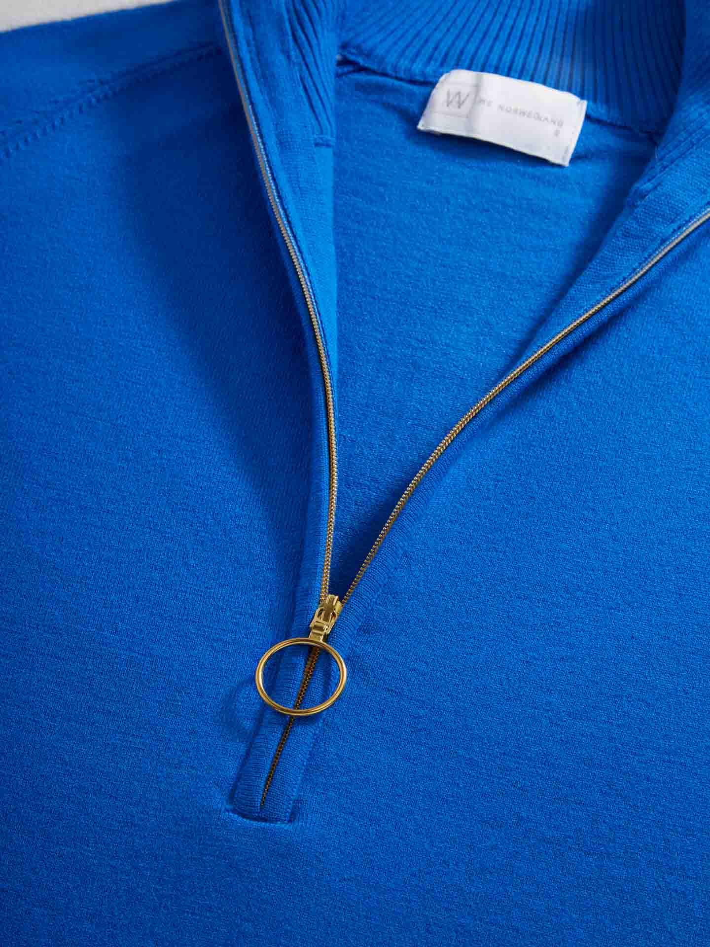 Close-up image of the Varden Zip Up Women, a blue half-zip women's ski sweater by We Norwegians. This 70s inspired sweater features a metallic zipper with a circular pull tab and a visible tag at the neckline. The texture of the fabric is smooth and finely knit, ideal for those chilly slopes.