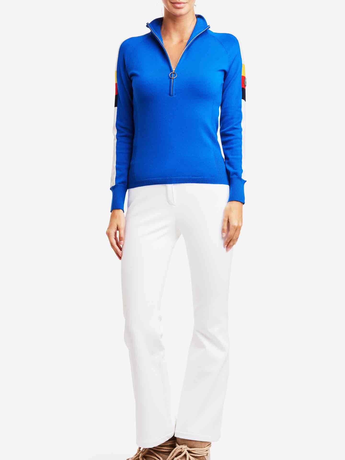 A person stands modeling a stylish outfit, featuring the Varden Zip Up Women by We Norwegians, a 70s inspired vivid blue, long-sleeve, half-zip ski sweater with multicolored patches on the sleeves. They pair it with white pants and tan footwear. The background is plain white.