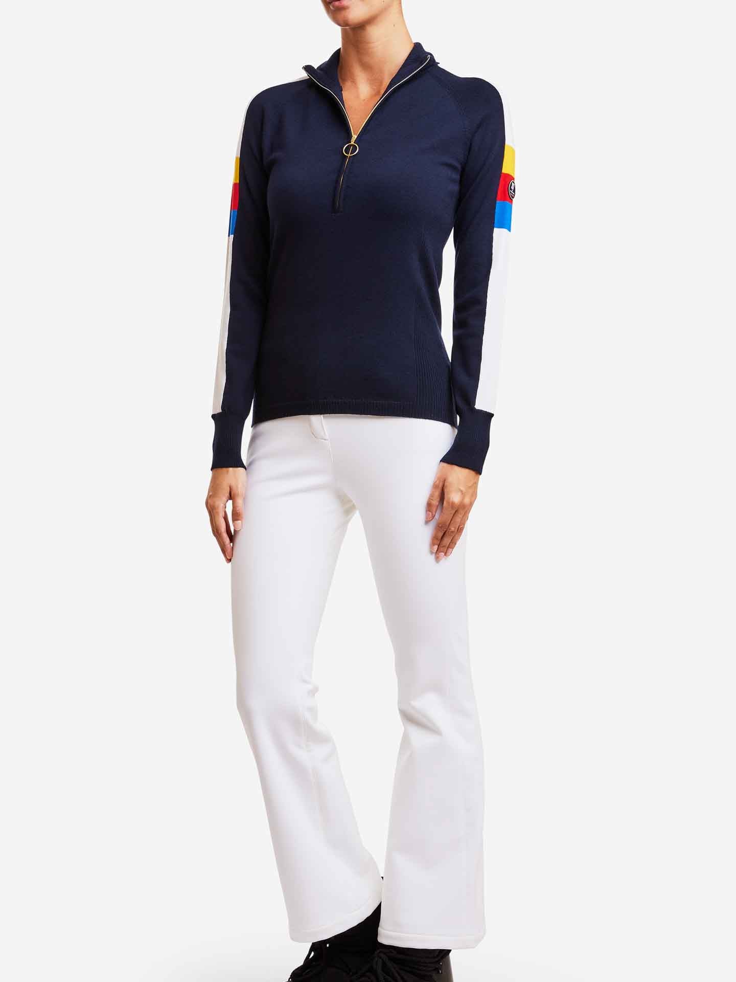 A person is wearing the We Norwegians Varden Zip Up Women, a navy blue women's ski sweater with a half-zip and colorful stripes on the sleeves, paired with white pants and black shoes, standing against a plain white background.