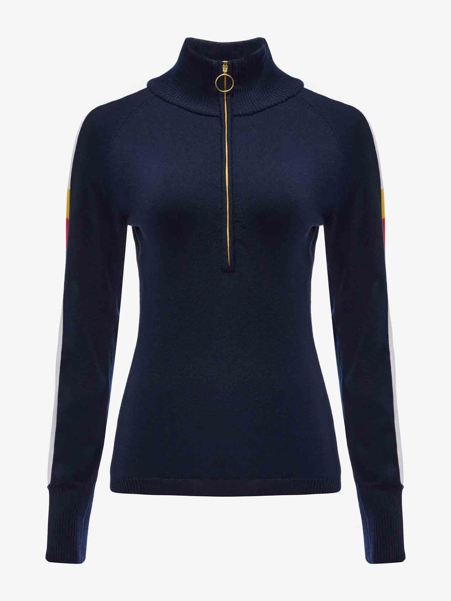 The Varden Zip Up Women by We Norwegians is a navy blue women’s ski sweater featuring a high collar and a gold zipper down the front. Made from Merino wool, the sweater has long sleeves adorned with white and red stripes, evoking a 70s inspired ski style. It is displayed against a white background.