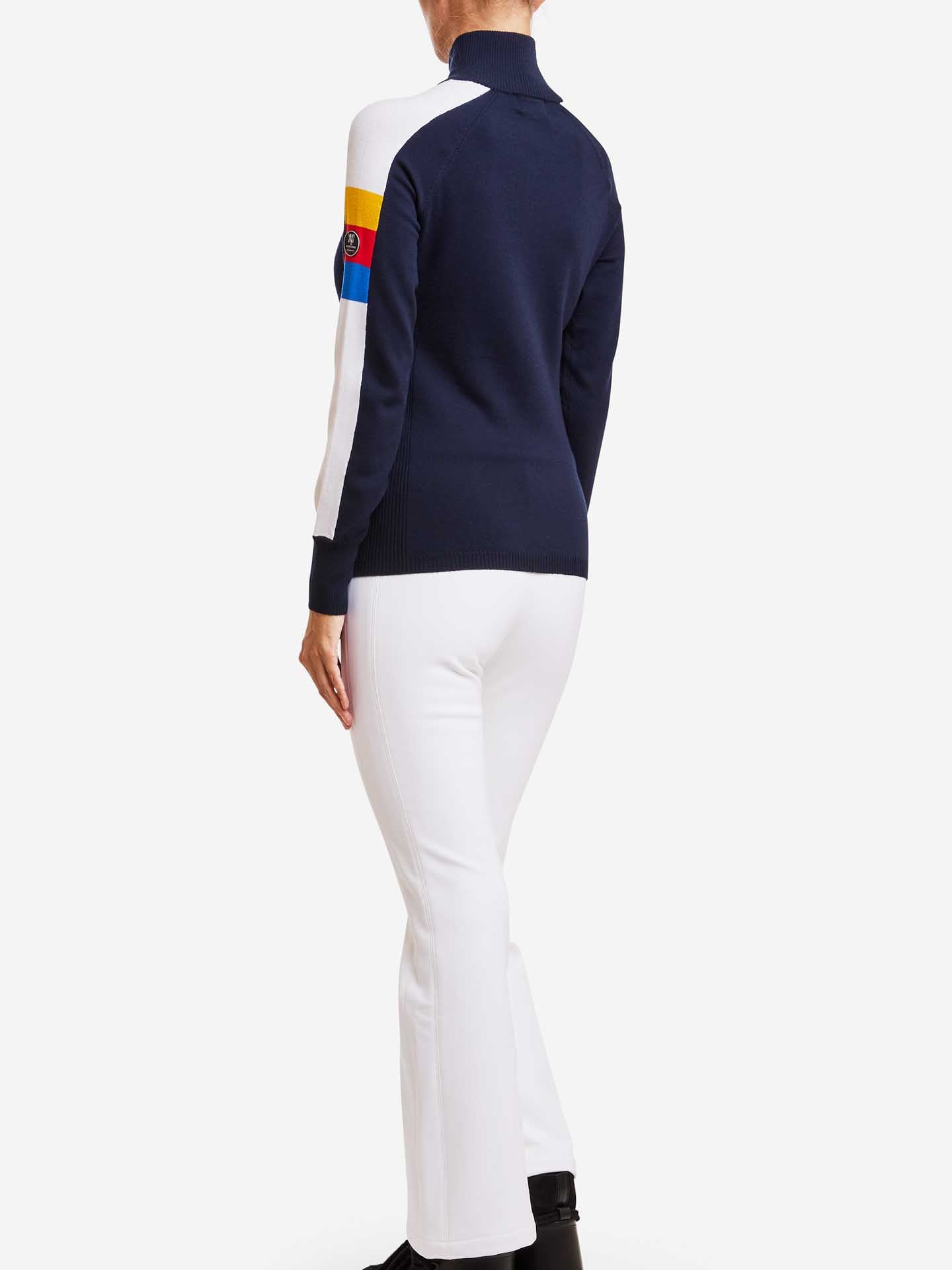 A person is shown from the back wearing the Varden Zip Up Women jacket in navy blue and white by We Norwegians, featuring multicolored patches on the sleeve. They are paired with white pants and black shoes, evoking a 70s-inspired ski sweater vibe as they stand against a plain white background.