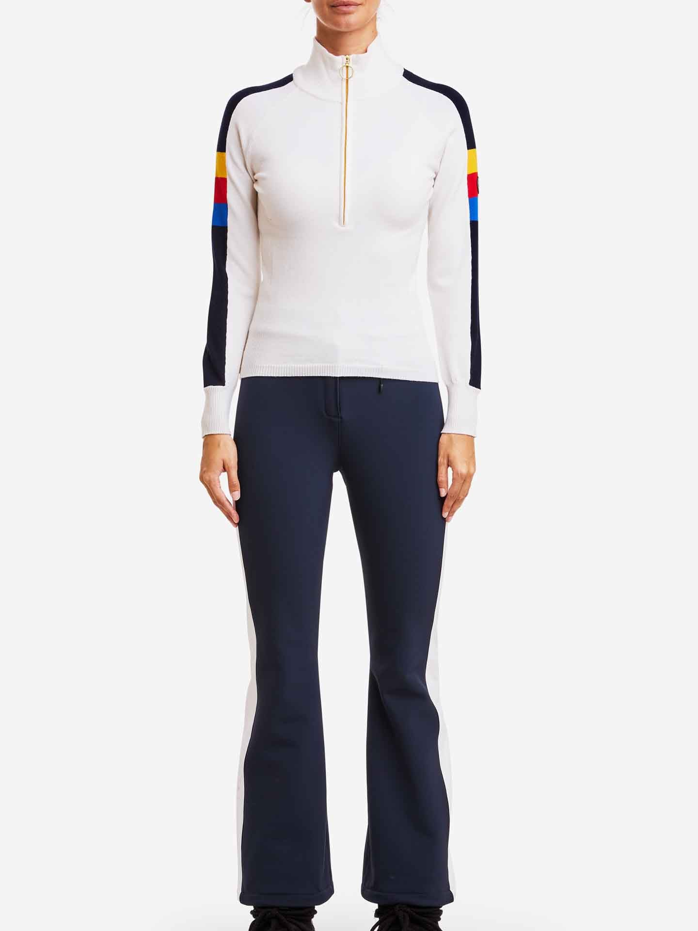 A person is wearing the "Varden Zip Up Women" sweater by We Norwegians, featuring a 70s inspired design with colored stripes on the sleeves and a zipper. They are also wearing dark navy pants with white side panels. The background is plain white.