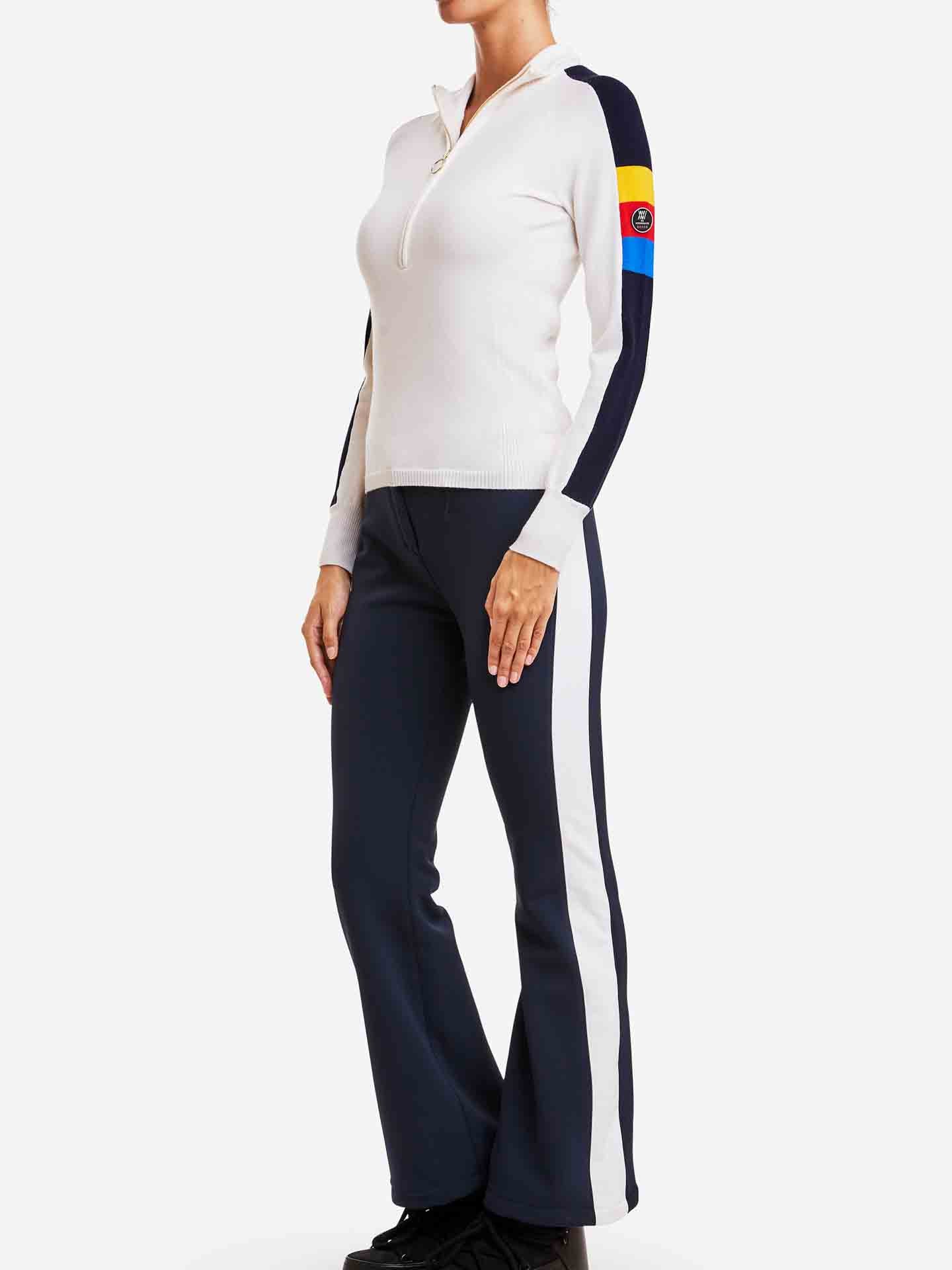 A person stands wearing the We Norwegians Varden Zip Up Women, a white long-sleeved top featuring black side stripes and colorful flag-like patches on the upper sleeve. They are also wearing black pants with white side stripes and black shoes, creating a look reminiscent of a 70s inspired women's ski sweater. The background is plain white.