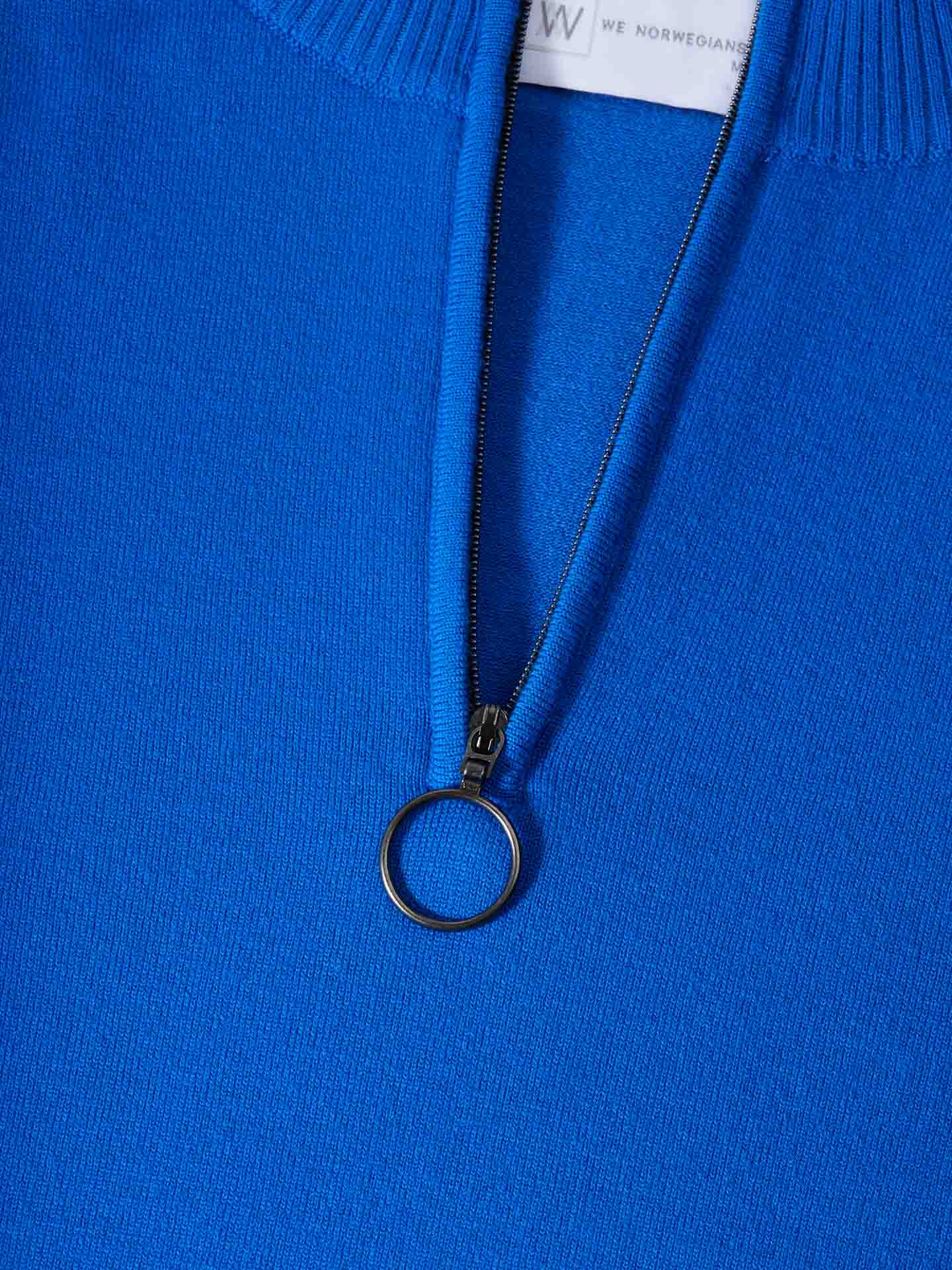 Close-up of a vibrant blue Merino wool sweater from We Norwegians, named the Varden Zip Up Men, featuring a quarter-zip design. The zipper is partially open, revealing a white tag with some text. The zipper pull consists of a small circular ring, adding a touch of elegance to the garment. This exquisite ski sweater is proudly made in Italy.