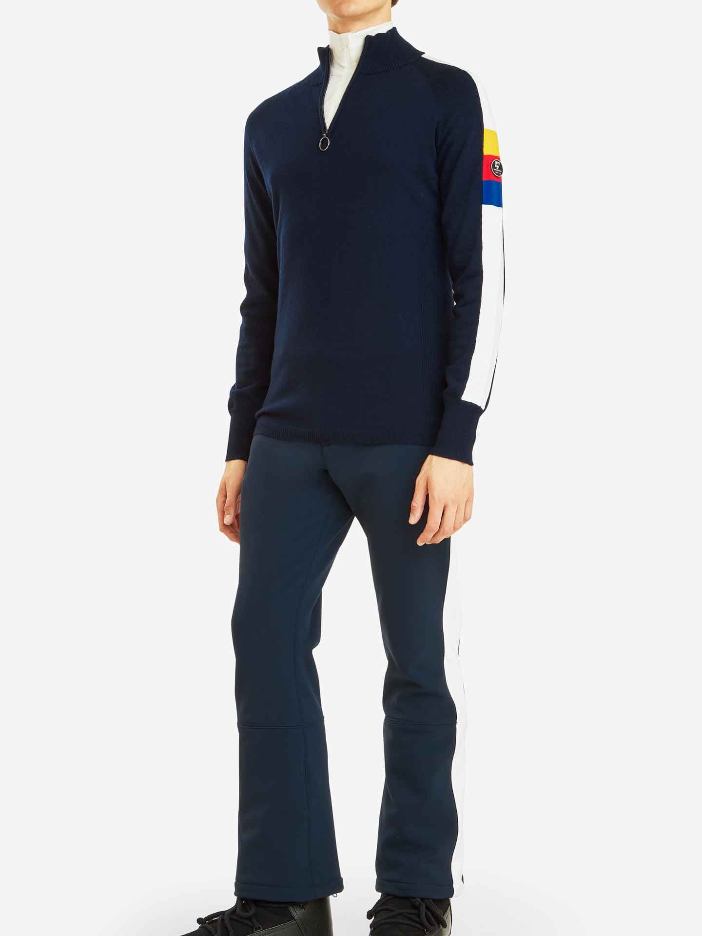 A person is wearing the We Norwegians' Varden Zip Up Men pullover in navy blue, which features a half-zip front and white stripes on the sleeves, paired with matching navy blue athletic pants with white side stripes. The pullover, reminiscent of a ski sweater, has a colorful patch on the upper arm. The background is plain white.