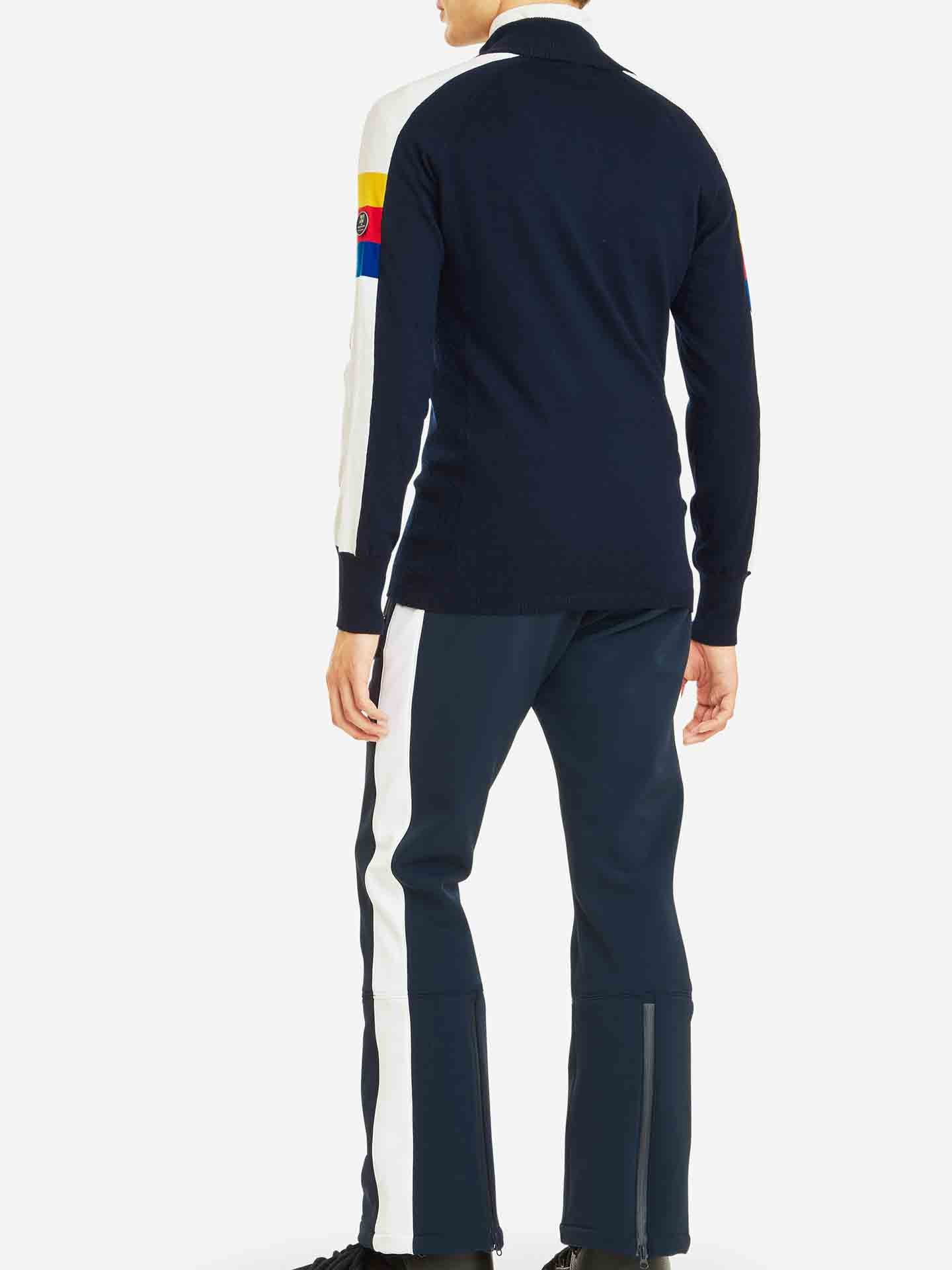 A man stands with his back to the camera, dressed in the "Varden Zip Up Men" ski sweater from We Norwegians, crafted in Italy. The dark ski sweater features white sleeves with colored bands near the wrists. He pairs it with navy blue pants accented by white stripes, and his right foot is slightly stepping forward.