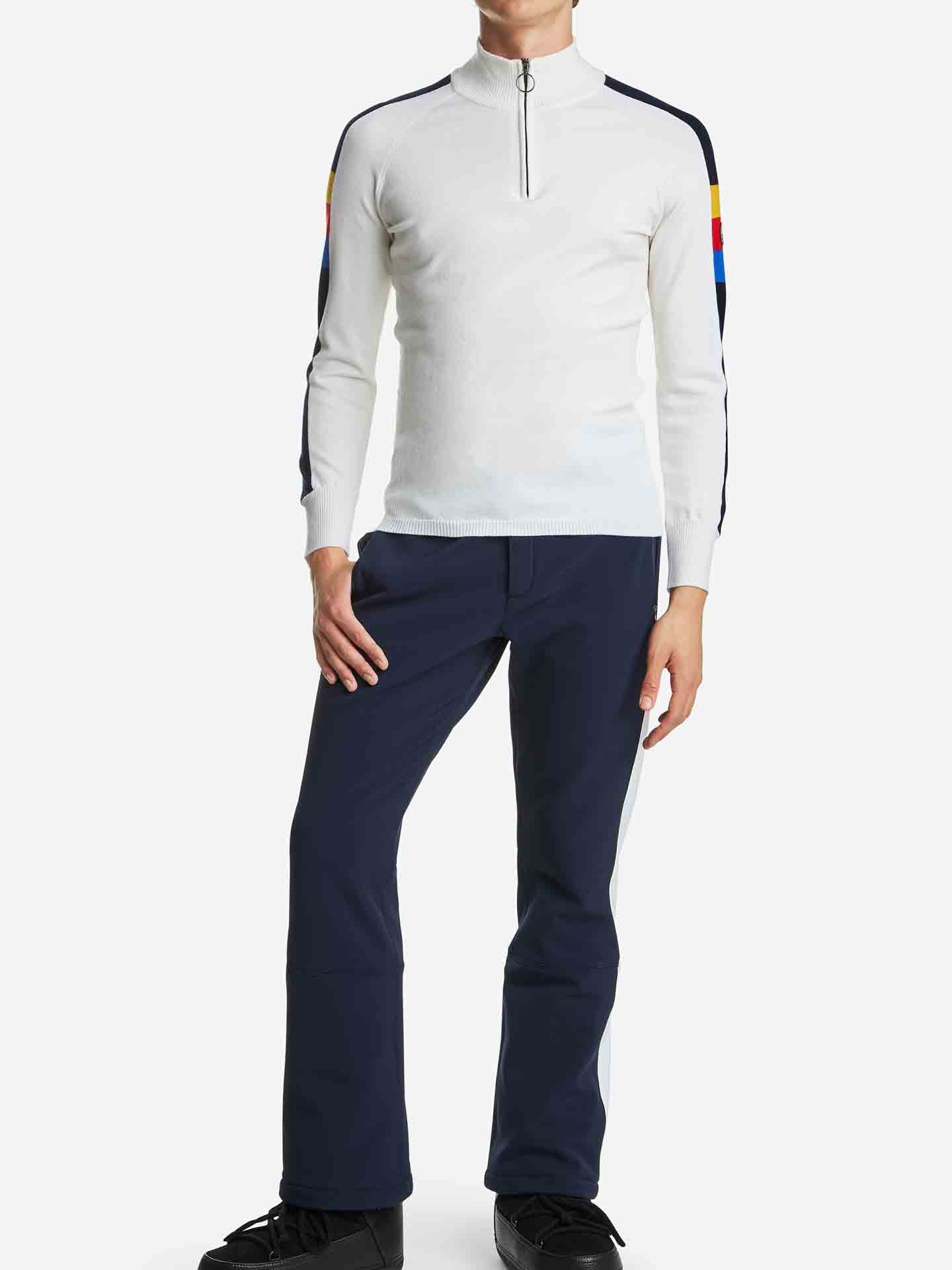 A person stands against a plain white background wearing the Varden Zip Up Men by We Norwegians, a white long-sleeve ski sweater featuring colorful stripes on the upper arms. Made in Italy from soft Merino wool, it pairs perfectly with their navy blue pants and black shoes, their right hand casually resting in their pocket.