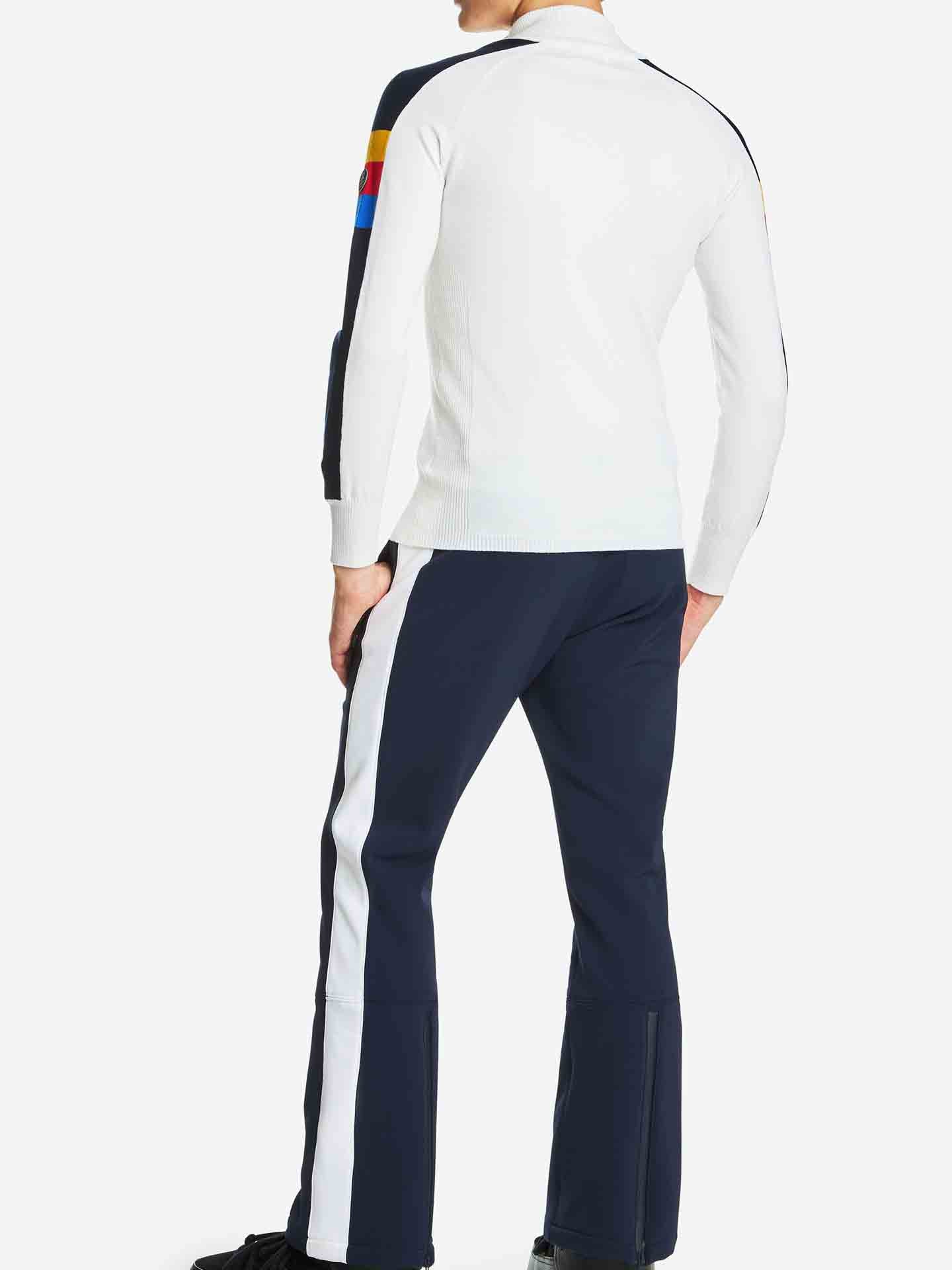 A man faces away from the camera, dressed in the Varden Zip Up Men jacket by We Norwegians, a white long-sleeve with colored stripes on the sleeves. He pairs it with navy blue pants featuring white stripes down the sides, evoking the style of a classic ski sweater. The background is white.