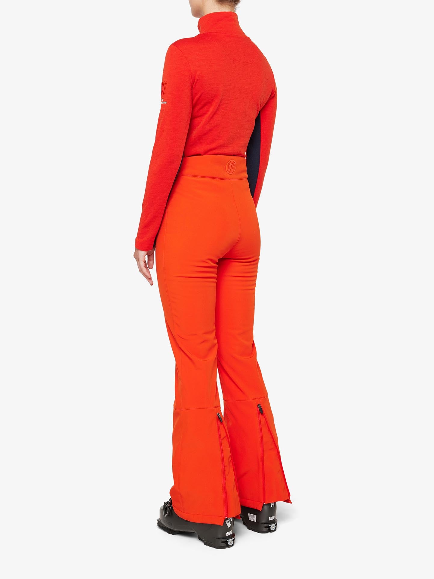 A person is sporting a vibrant orange ski ensemble featuring the 'Tyin Bootleg Ski Pant Women' from We Norwegians, complete with flared pants adorned with zippers at the back and a matching long-sleeve top. Their black ski boots are enhanced by snow gaitors. The individual stands facing away from the camera against a white background.