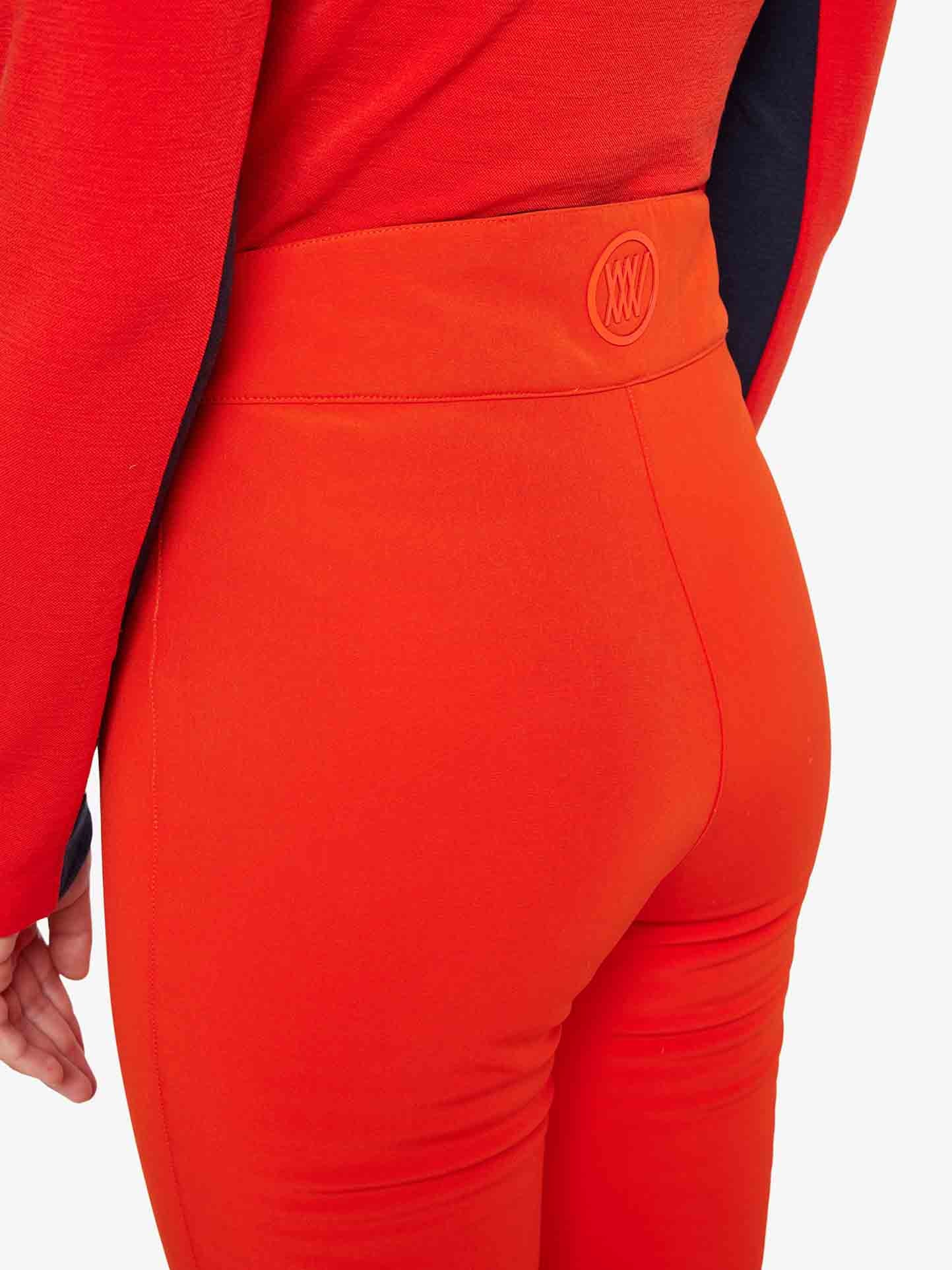 A person is wearing the bright red Tyin Bootleg Ski Pant for Women by We Norwegians, featuring a symbol on the high waistband, reminiscent of upscale ski sweaters. Their red top peeks out slightly, and their left arm is bent at the elbow against a plain white background.