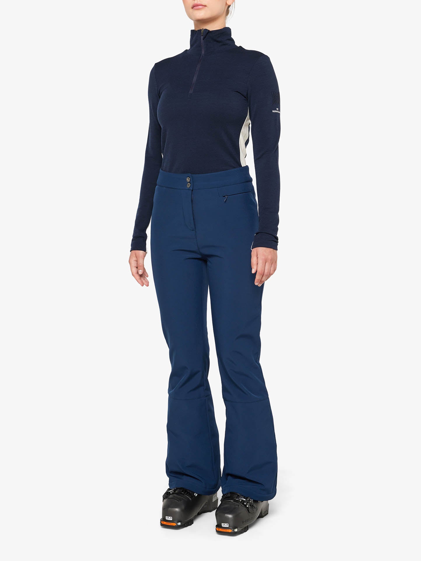 A person stands against a white background, clad in a navy blue long-sleeved, zip-up top and We Norwegians' Tyin Bootleg Ski Pant for women, paired with black ski boots. The water-repellent soft shell attire hints at readiness for snow adventures. Their face is not visible in the image, and their hands are relaxed at their sides.