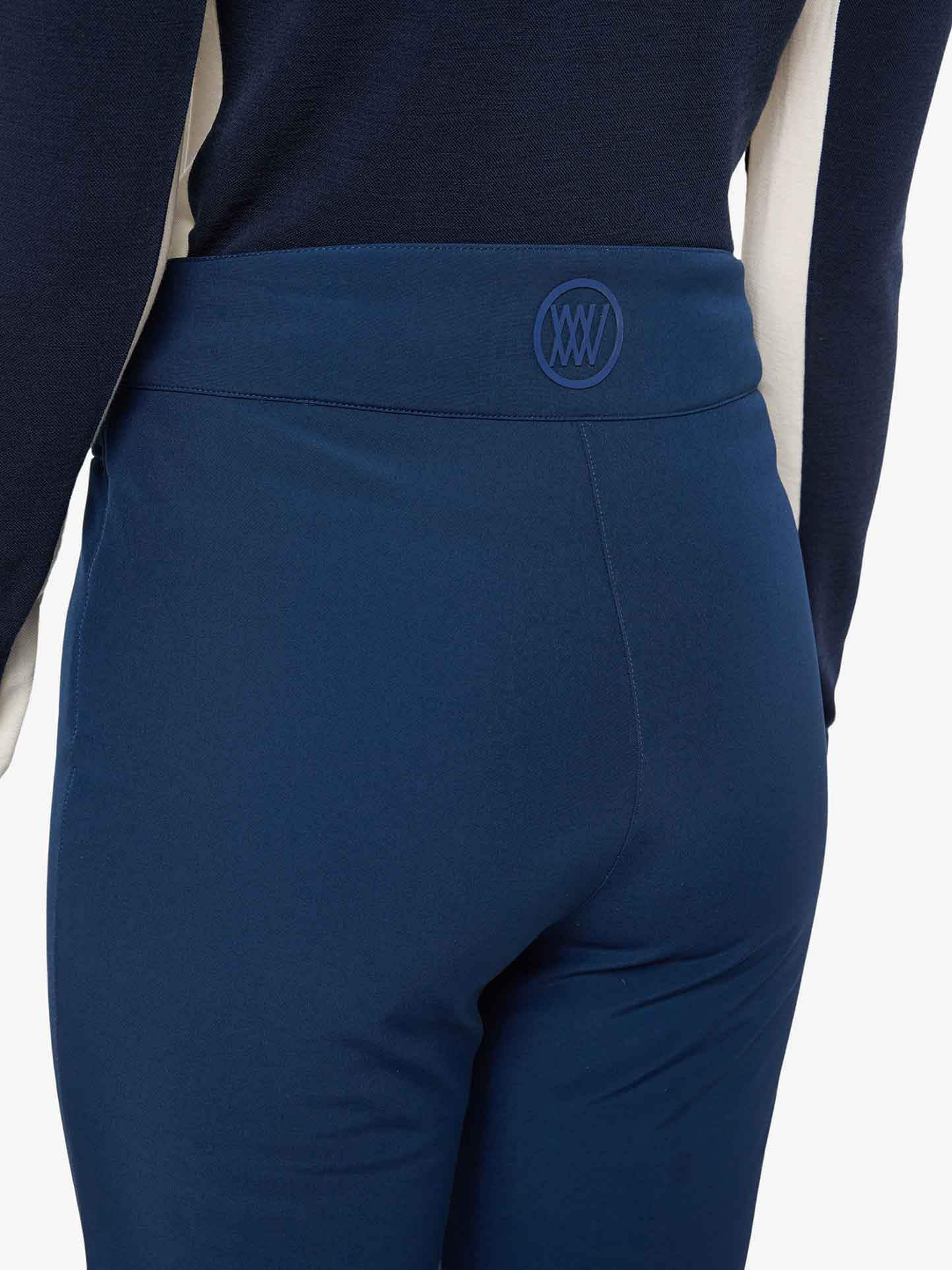 A person is seen from the back, wearing blue pants and a black and white top. The waistband of the **Tyin Bootleg Ski Pant Women** by **We Norwegians**, made from water repellent soft shell material, features a circular logo with overlapping lines in the center. The image focuses on the lower back and pants.