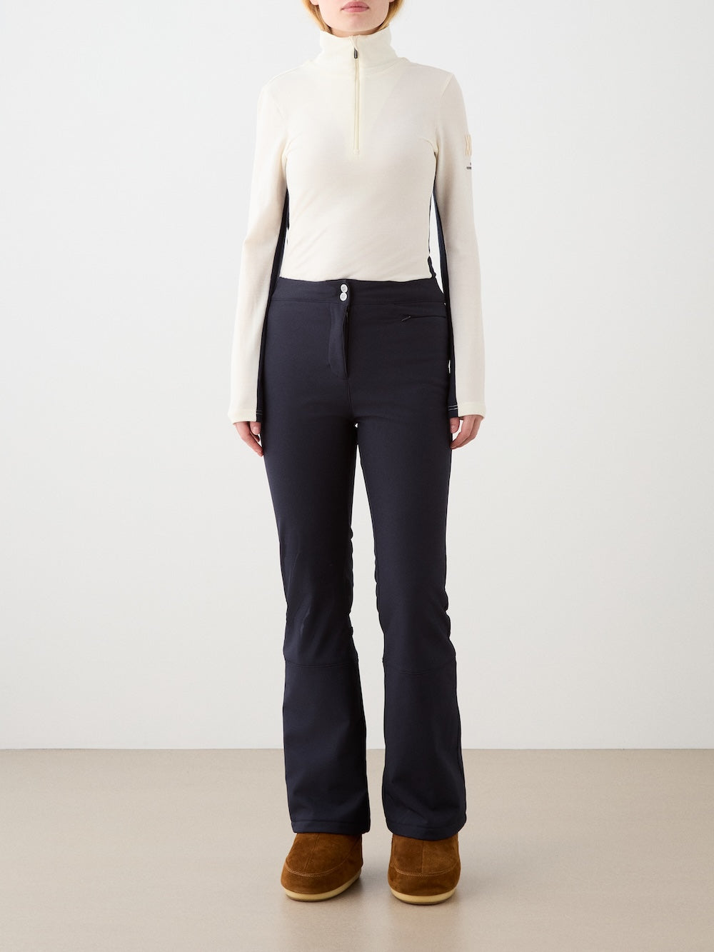 A woman faces forward against a plain background, wearing a cream ski sweater, brown boots, and We Norwegians Tyin Bootleg Ski Pant Women.