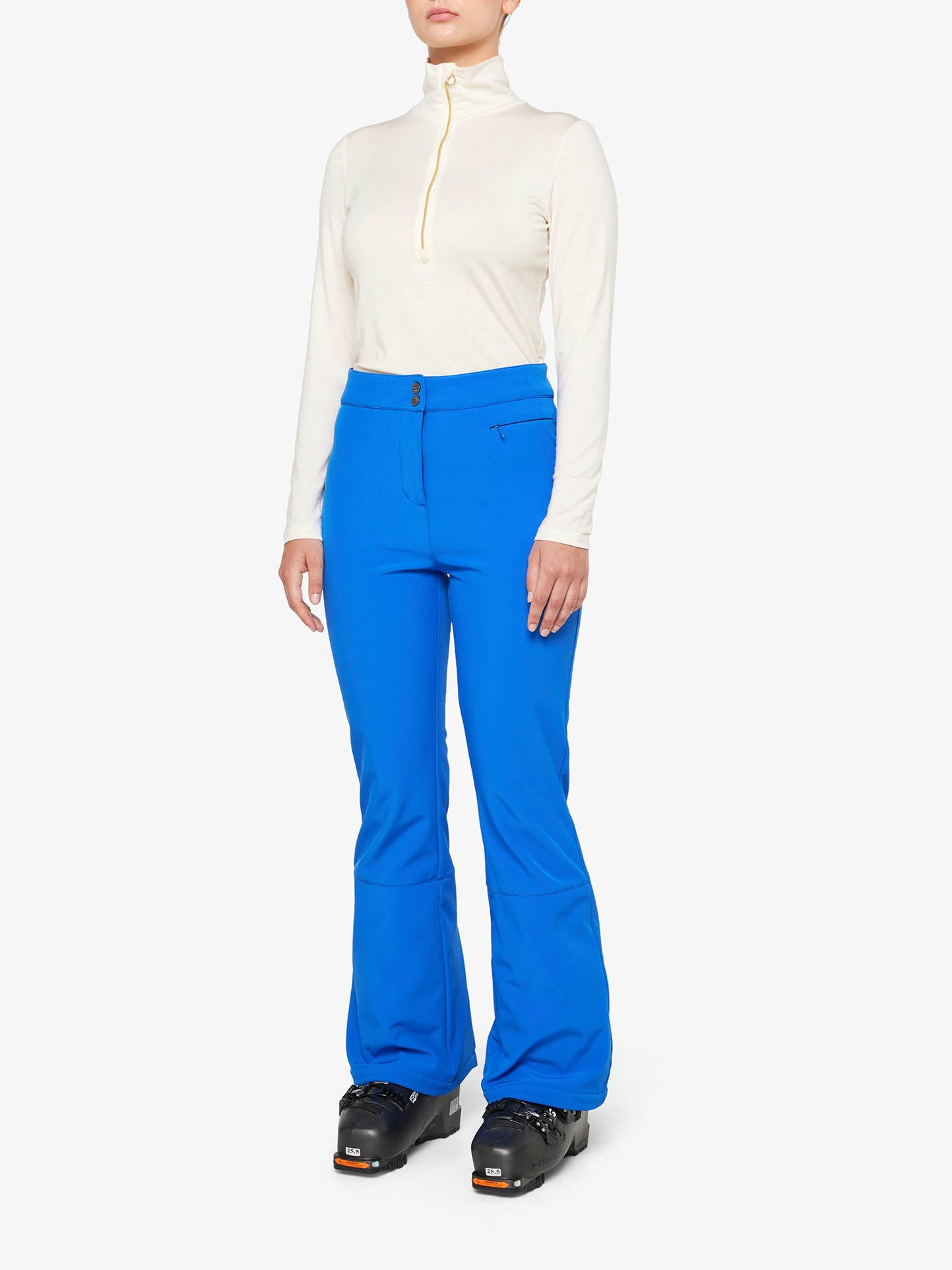 A person is dressed in a white, long-sleeve turtleneck and the bright blue *We Norwegians Tyin Bootleg Ski Pant Women* with snow gaitors, standing upright. They are also wearing black ski boots. The background is plain white.