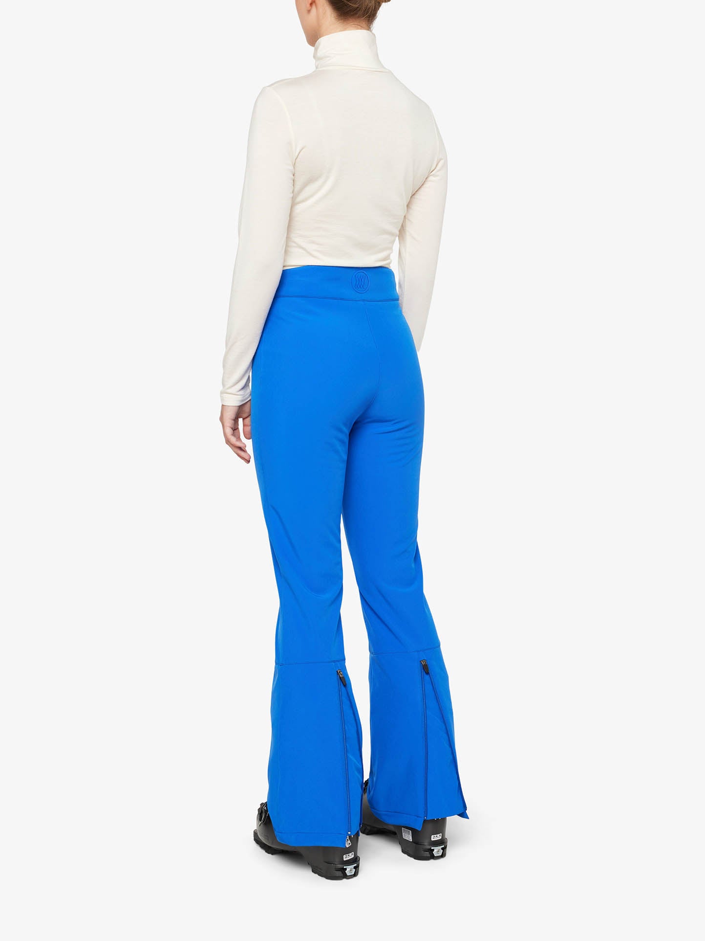 A person is shown from behind, wearing a white turtleneck top and bright blue We Norwegians Tyin Bootleg Ski Pant Women. The pants, complete with zippered flares at the bottom and black ski boots, feature integrated snow gaitors. The background is plain white.