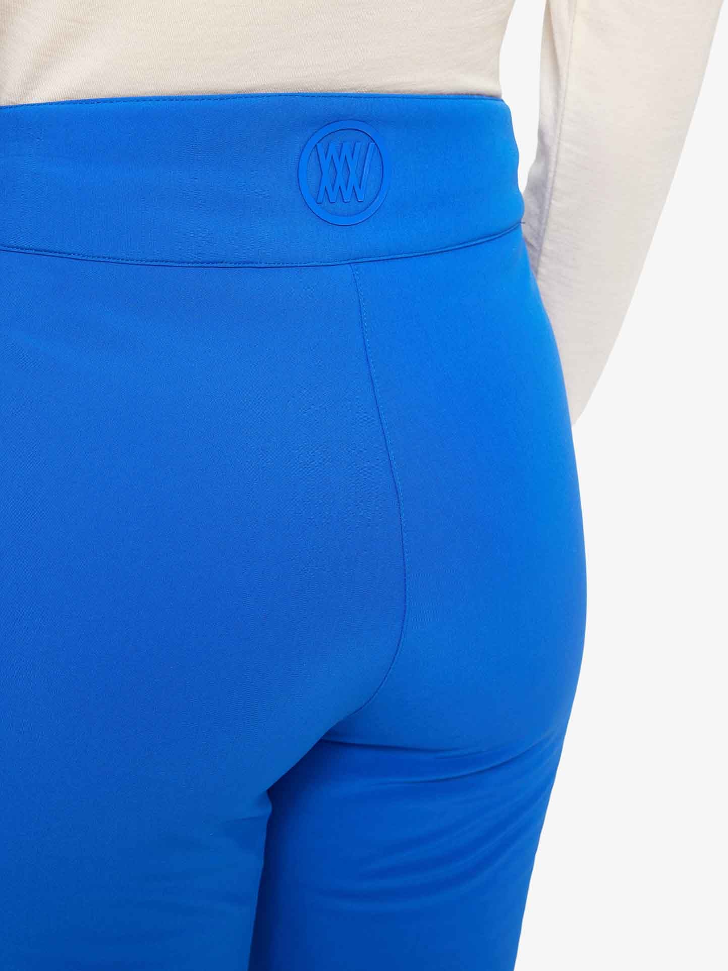 A person wearing the bright blue Tyin Bootleg Ski Pant Women by We Norwegians and a white top is shown from the back. The waistband of the water-repellent soft shell pants features an embossed round logo with an intricate design. The person’s upper body and the lower part of the pants are partially visible.