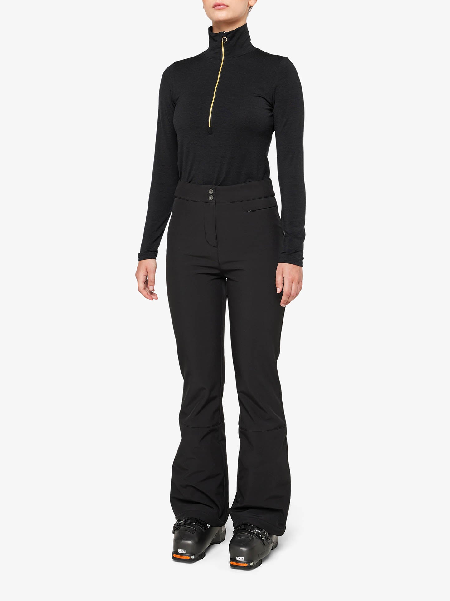 A person stands against a plain background wearing a sleek, black long-sleeve zip-up top made of soft shell fabric and the We Norwegians' Tyin Bootleg Ski Pant Women in matching black. Their hands rest at their sides, and they also wear water-repellent ski boots. The outfit appears stylish and fitted, perfect for cold-weather activities.