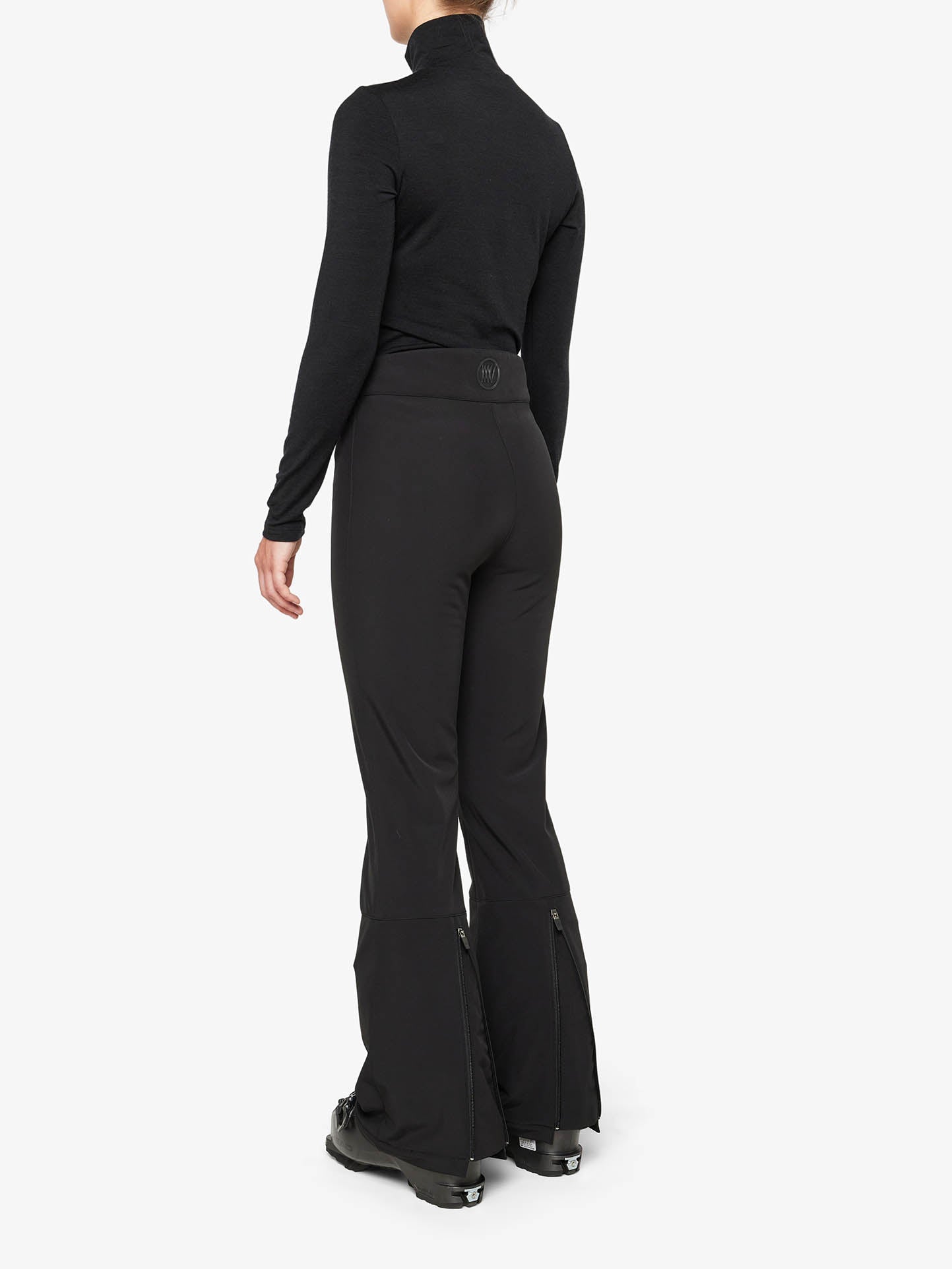 A person is seen from behind wearing a black turtleneck and the Tyin Bootleg Ski Pant for women by We Norwegians, featuring flared bottoms with zippers. The water repellent soft shell pants are paired with matching black shoes, and the individual is standing against a plain white background.