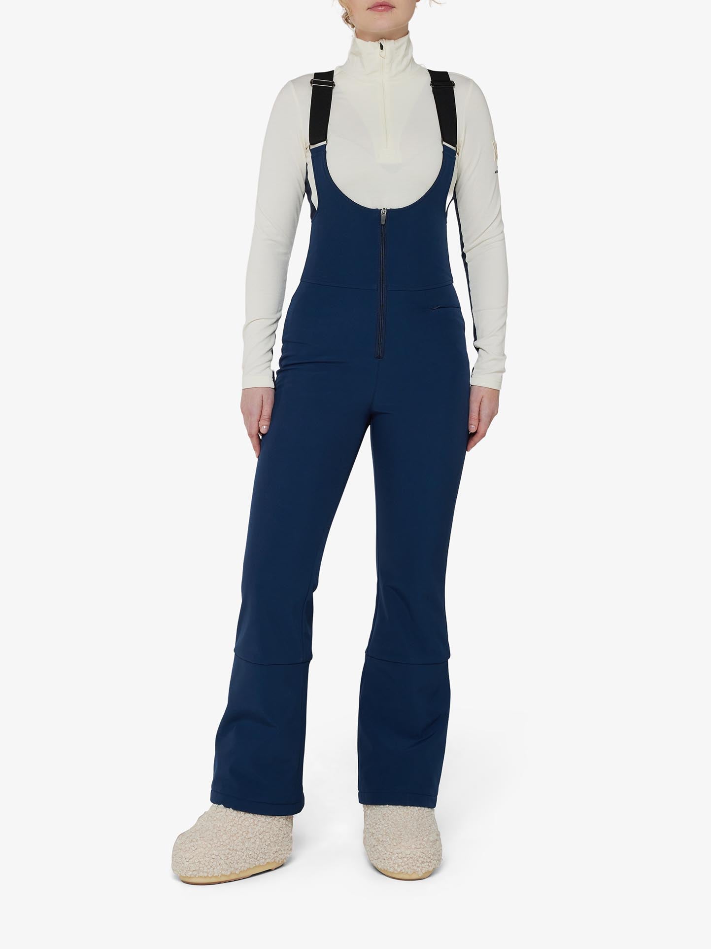 A person is wearing a cream long-sleeve shirt under the Tyin Bib Pant Women by We Norwegians, which features black straps. Made of water-repellent soft shell fabric, the navy blue, high-waisted design provides both style and functionality. They are also wearing beige, fluffy slip-on shoes and standing against a white background, highlighting a cozy outfit perfect for cold weather.