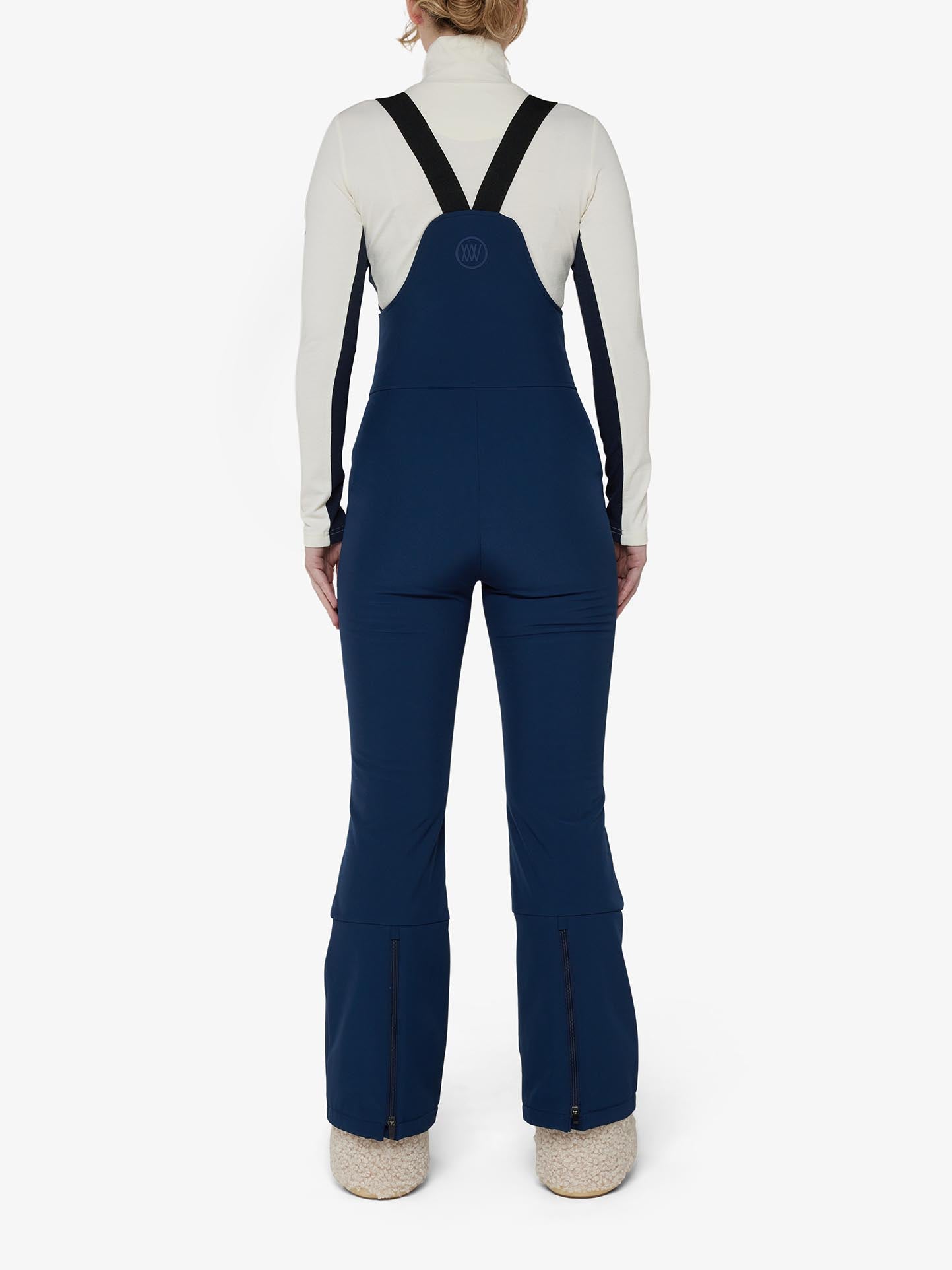 Revised Sentence: Rear view of a person wearing a white long-sleeve top paired with We Norwegians' Tyin Bib Pant Women in navy blue, featuring black shoulder straps. These high-waisted overalls showcase wide-leg bottoms with zippers at the ankles and are made from water-repellent soft shell fabric. The person stands against a plain white background.