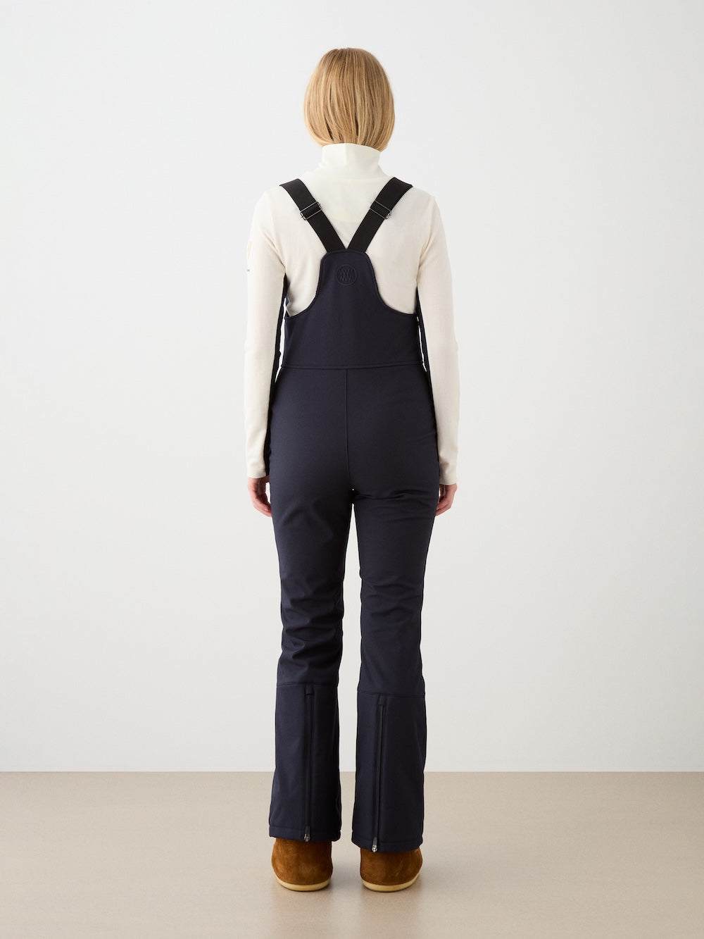 A person with short blonde hair stands indoors against a plain, light-colored background, facing away while wearing a white turtleneck and the We Norwegians Tyin Bib Pant Women—dark water-repellent soft shell overalls with black straps designed for superior alpine performance. They complete the look with brown shoes.
