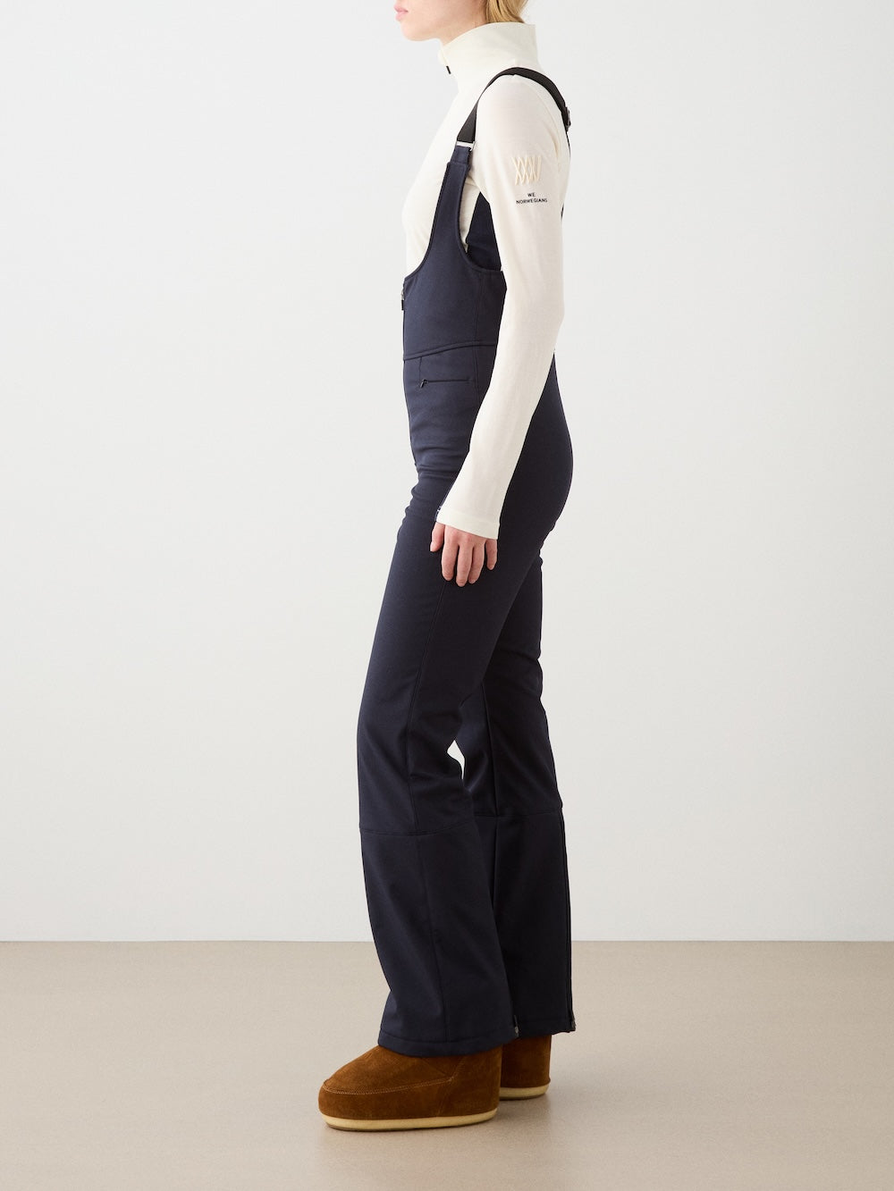 A person in a cream turtleneck top and black We Norwegians Tyin Bib Pant Women overalls stands sideways, wearing brown suede slip-on boots in a minimal indoor setting with a plain background—blending everyday style with alpine performance.