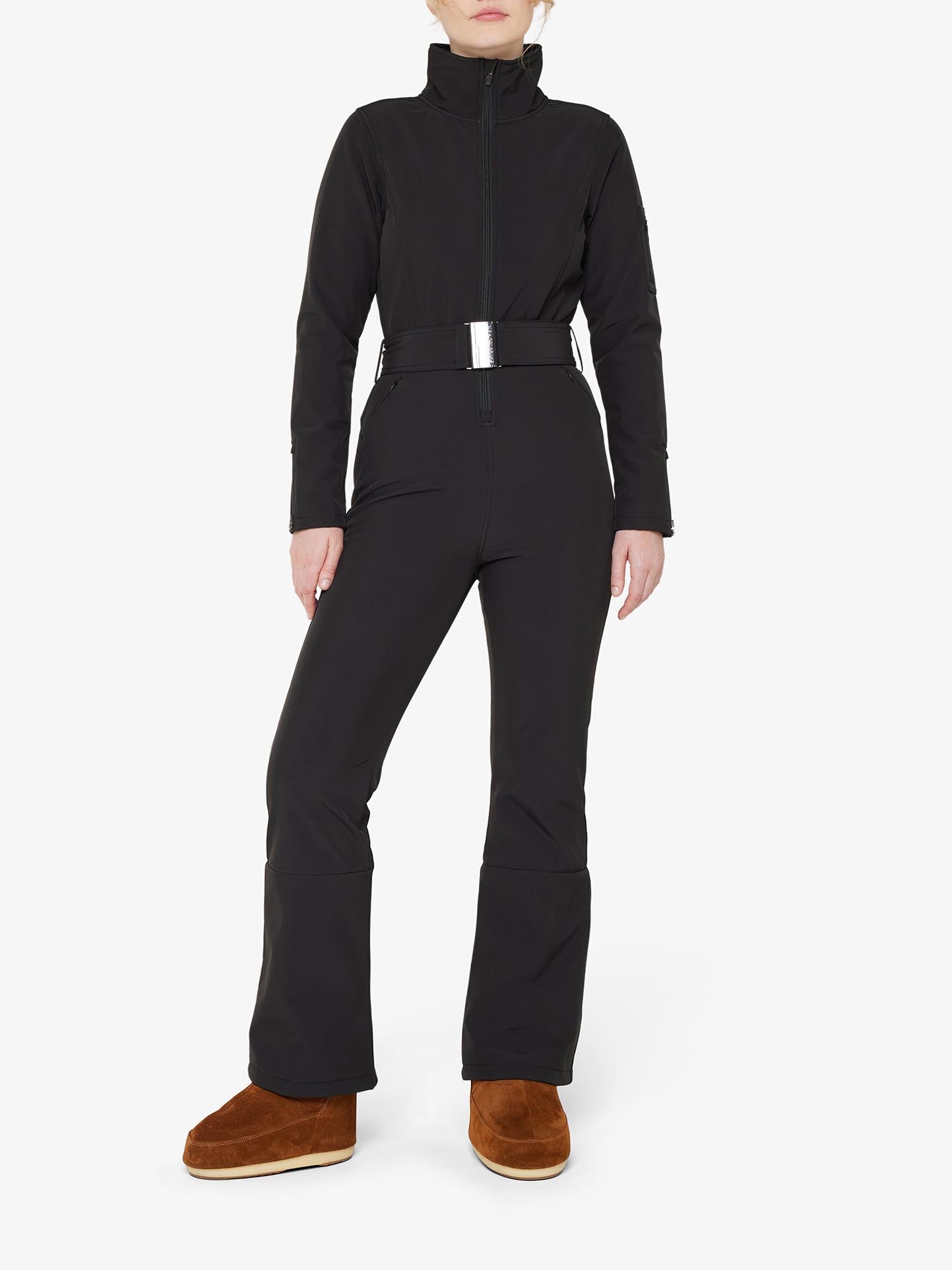 A person wearing the We Norwegians Tyin Ski Suit Women, a black, water repellent jumpsuit with a high collar and belted waist, stands against a plain white background. The outfit is complemented by tan-colored, fur-lined boots and features convenient ski card pockets.