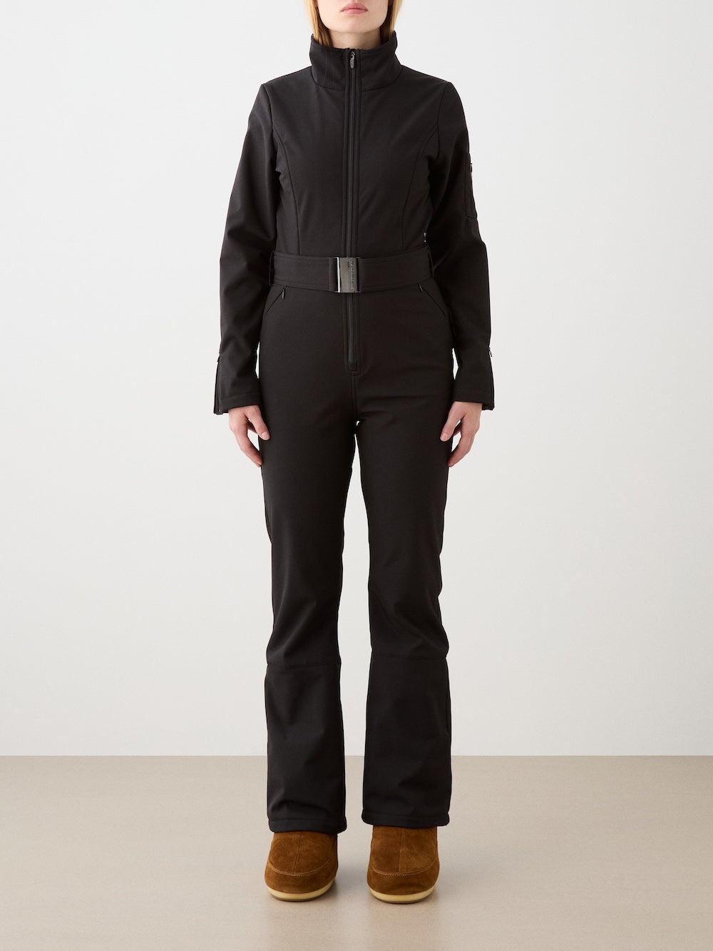 A woman faces forward in a fitted black We Norwegians Tyin Ski Suit Women—a water-repellent soft shell ski suit with high collar—paired with brown winter boots, standing against a plain light background.