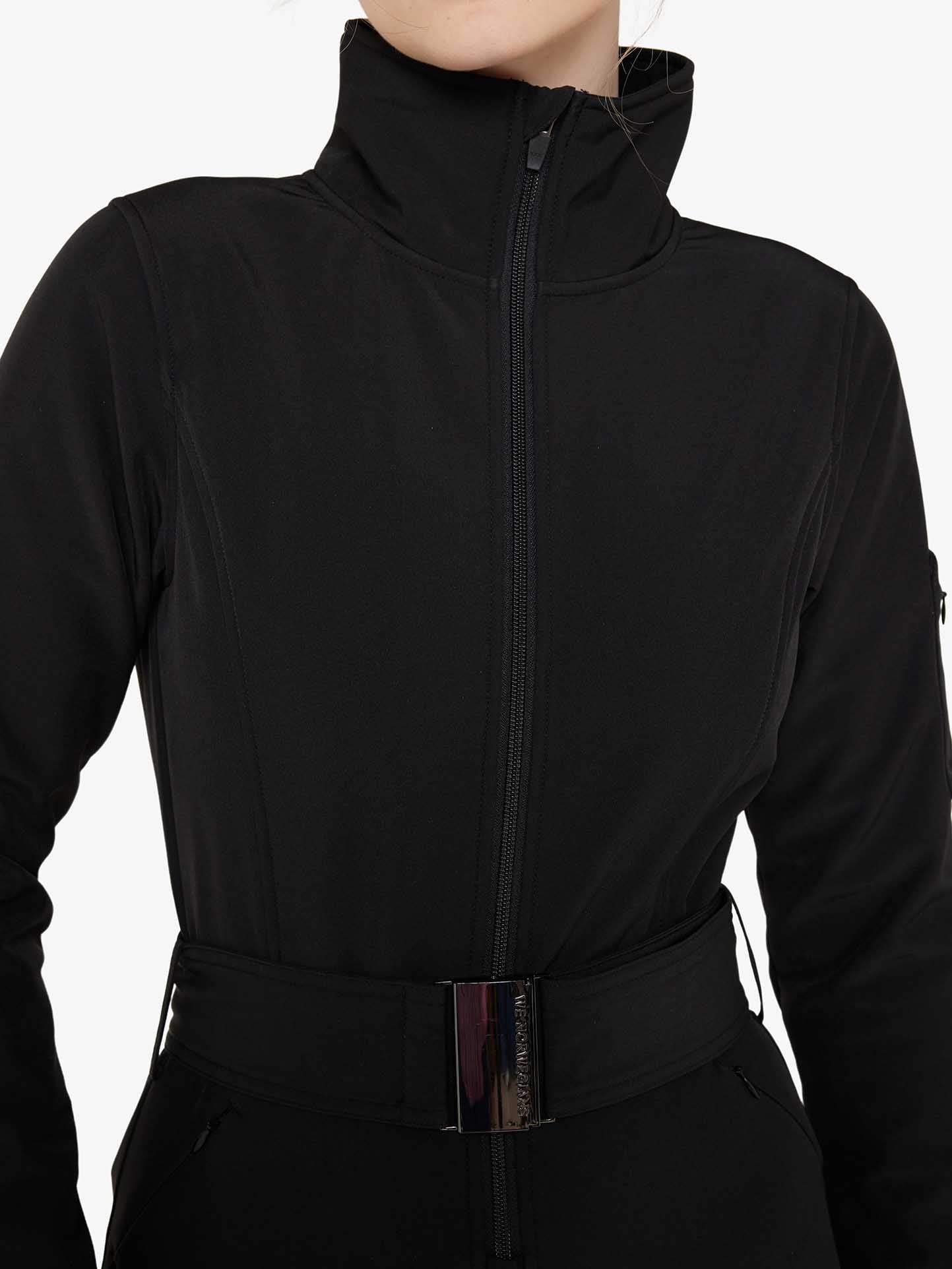 A person is wearing the Tyin Ski Suit Women by We Norwegians, a black long-sleeve jacket with a high neck and zipper closure, featuring a belt with a metallic buckle at the waist. The sleek design also includes a convenient ski card pocket. The background is plain white.