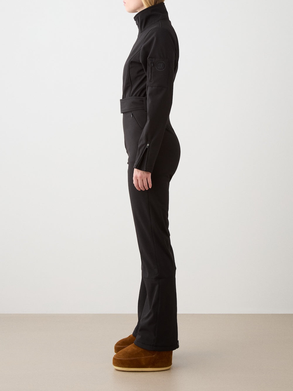 A woman stands in profile against a plain background, wearing the fitted We Norwegians Tyin Ski Suit Women—a water-repellent soft shell ski suit with a high collar and brown platform shoes.