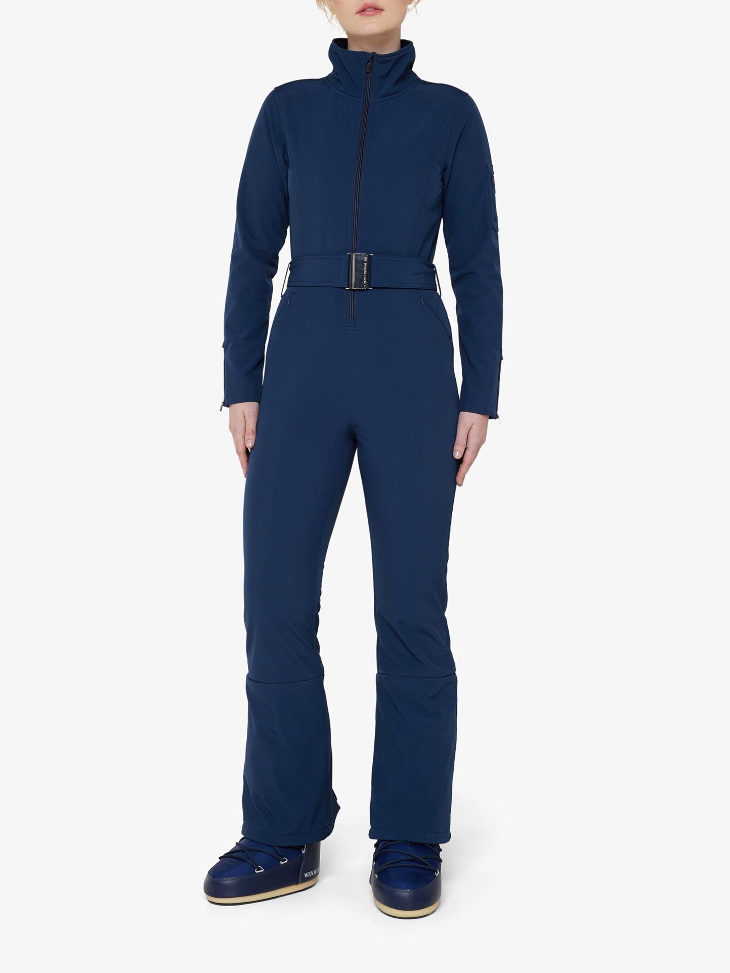 A person is wearing the Tyin Ski Suit Women by We Norwegians in navy blue. The water-repellent jumpsuit features a front zipper, high collar, and a belt around the waist. They are also sporting matching navy blue snow boots with white soles. The ski suit includes a convenient ski card pocket. The background is plain white.