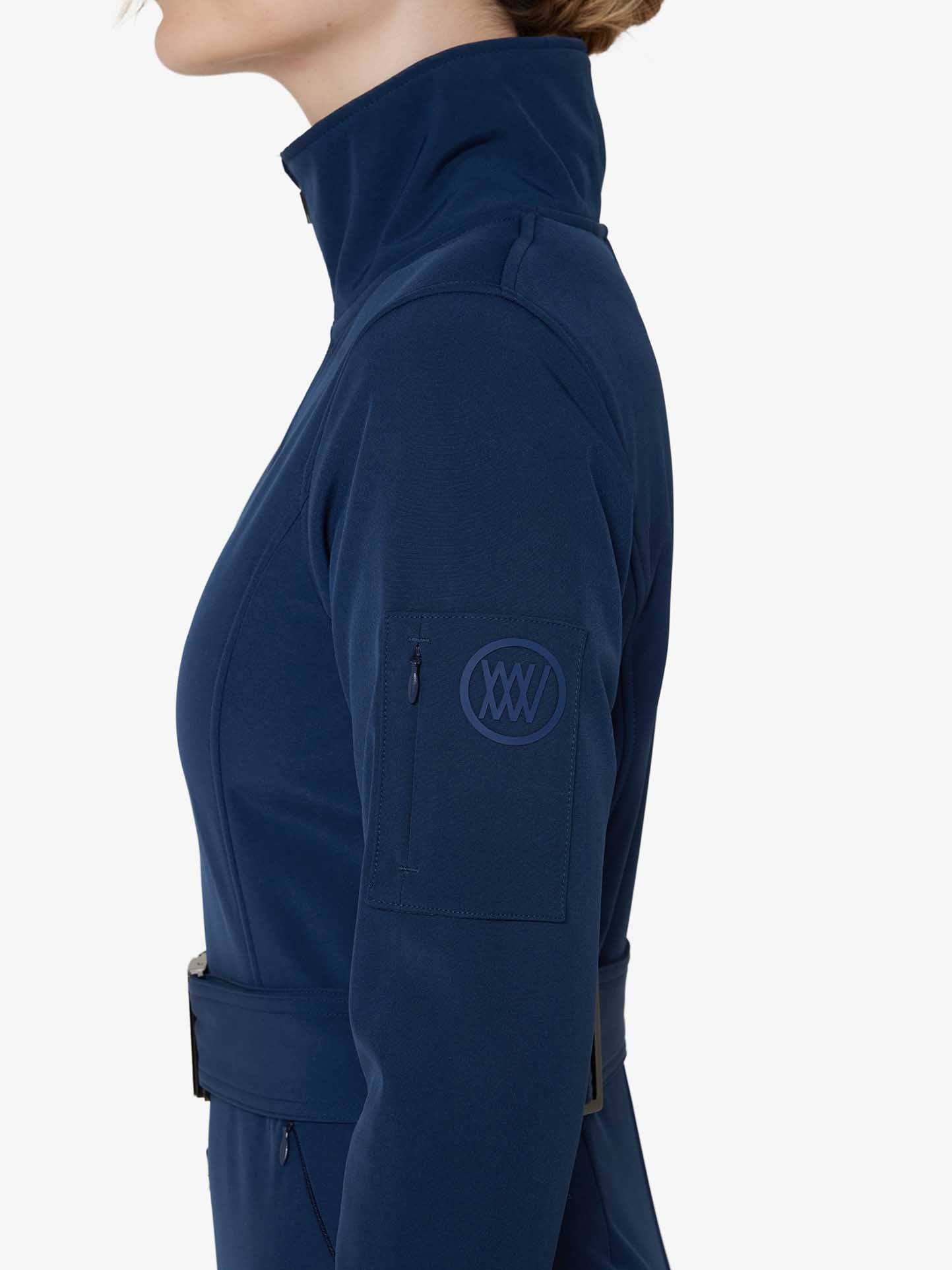 Side profile of a person wearing the We Norwegians Tyin Ski Suit Women in dark blue with a high collar. The ski suit, designed for water repellence, features a zippered pocket on the left sleeve that bears a circular logo. The individual’s face is not visible, and the background is plain white.