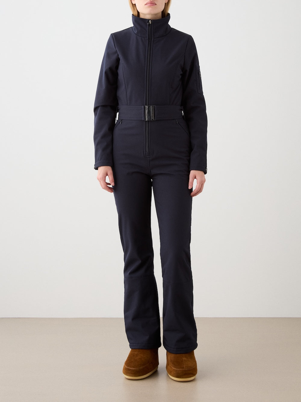 A person stands against a plain background wearing the fitted We Norwegians Tyin Ski Suit Women, a long-sleeve black ski suit in water-repellent soft shell with a high collar, belted waist, and brown boots. The face is not visible.