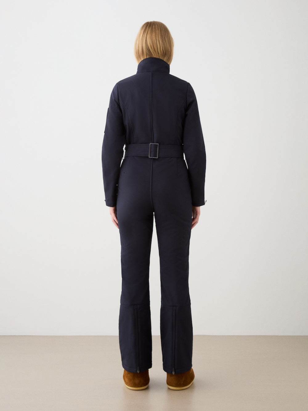 A woman with short blonde hair stands facing away, wearing the We Norwegians Tyin Ski Suit Women—a dark, water-repellent soft shell—paired with brown boots, against a plain light background.