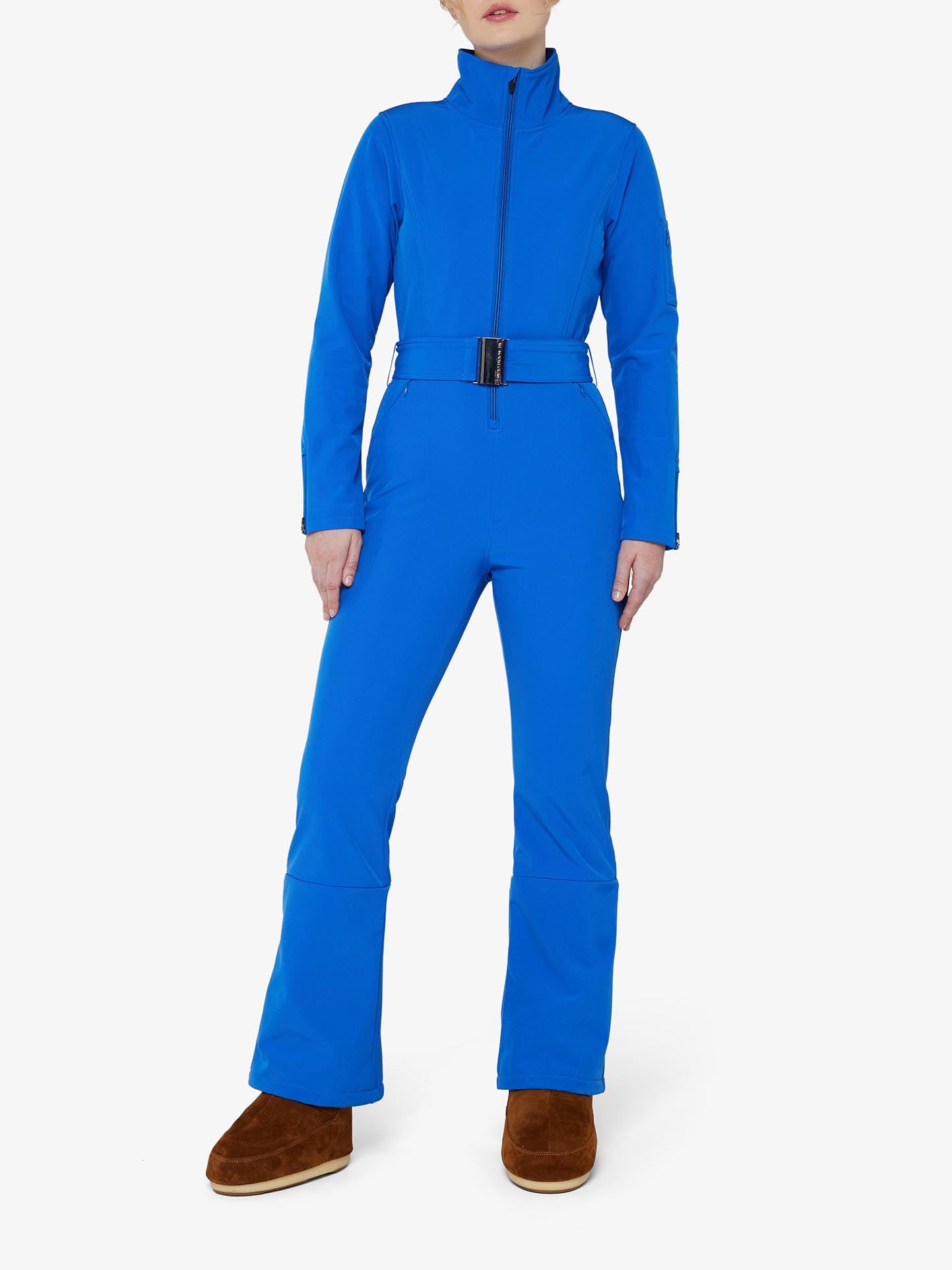 A person wearing the We Norwegians Tyin Ski Suit Women, a bright blue water-repellent jumpsuit with a high collar and belt, paired with brown boots. The Tyin Ski Suit features snow gaiters and a convenient ski card pocket, designed for winter or outdoor activities, providing full coverage and warmth. The background is plain white.