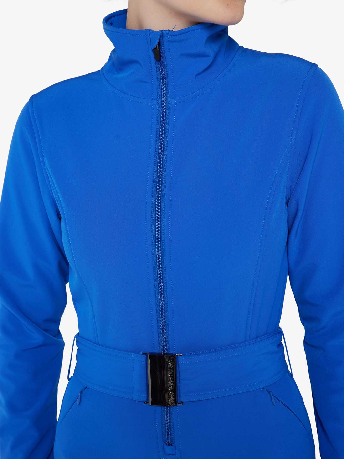 A person is wearing the Tyin Ski Suit Women by We Norwegians, characterized by a bright blue, zip-up jacket with a matching belt featuring a black buckle. The jacket includes a high collar, long sleeves, and convenient ski card pockets. The image is cropped to show only the torso, omitting the person's head and lower body.