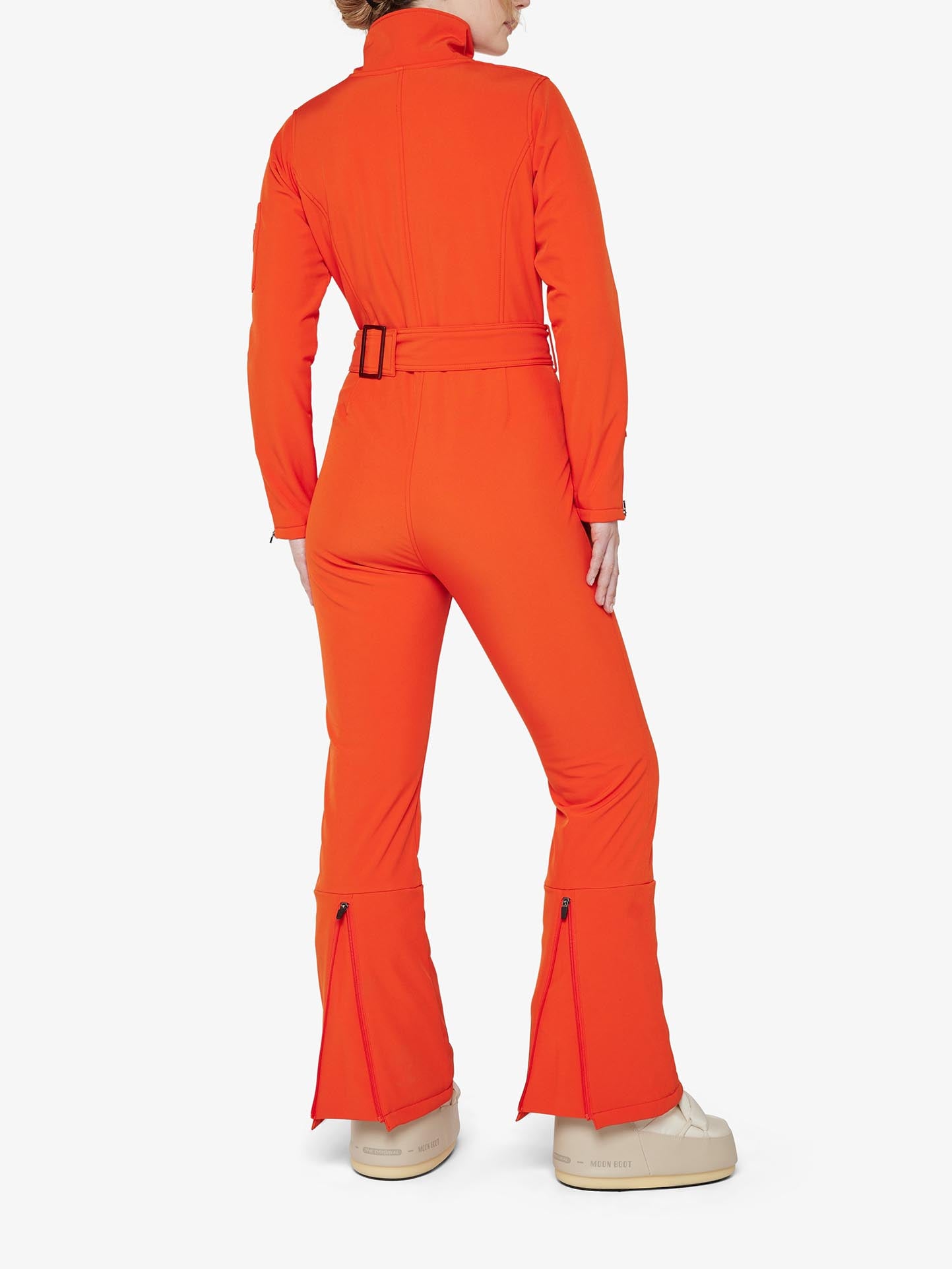 A woman wearing the Tyin Ski Suit Women by We Norwegians, an orange, water-repellent jumpsuit featuring a belted waist and flared legs with zippered vents at the bottom of each leg, posed with her back turned to the camera. She is also wearing white shoes and standing against a plain white background.