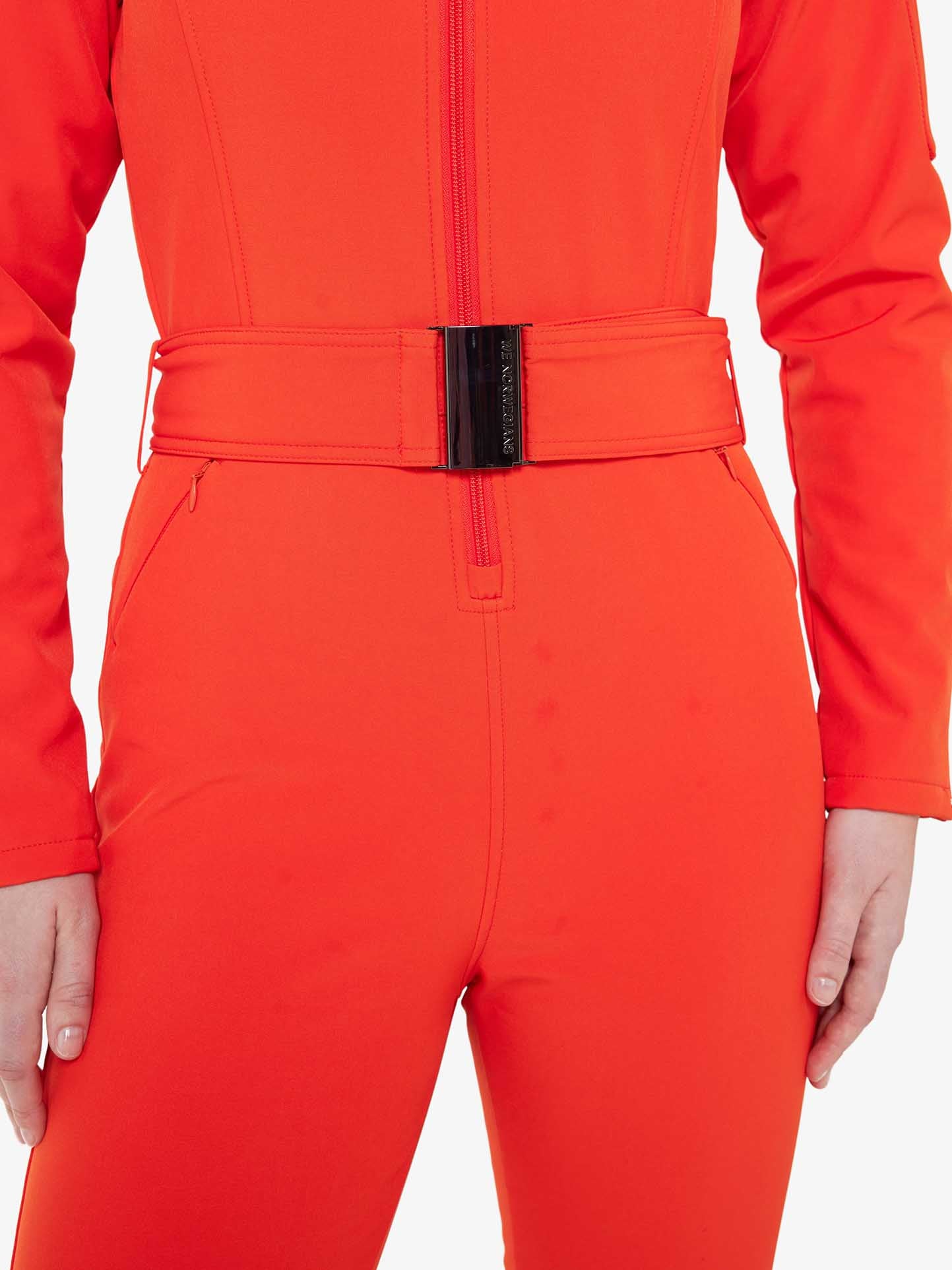 Close-up of a person wearing the Tyin Ski Suit Women by We Norwegians, a bright red, water-repellent jumpsuit complete with a matching belt featuring a black clasp. The person's hands are partially visible, resting near the waistband. The jumpsuit has a front zipper extending down to the belt and includes a convenient ski card pocket. The background is plain white.