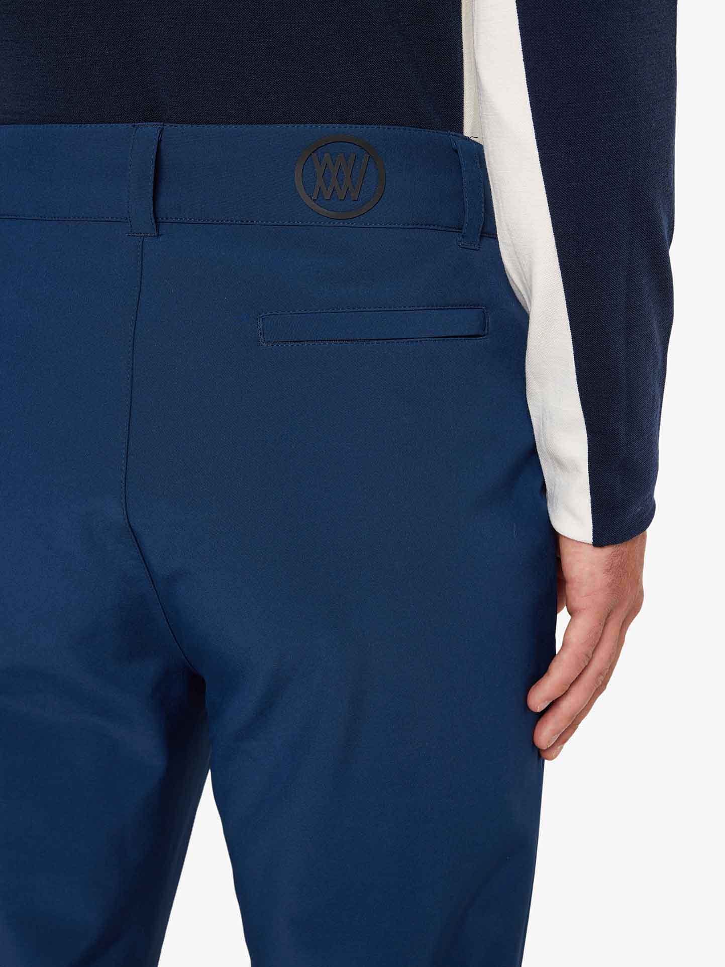 A person wearing a black and white long-sleeve shirt and the Tyin Ski Pant Men from We Norwegians, which are slim fit blue pants featuring a belt loop and a small embroidered logo on the waistband, is shown from the back. Their left hand is relaxed by their side.