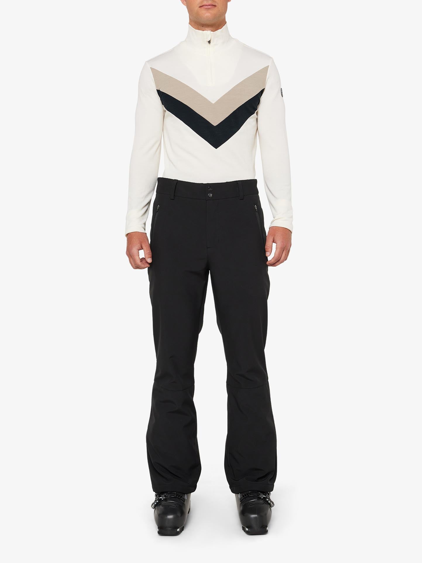 A person stands facing the camera wearing a white long-sleeve top with beige and black chevron designs, along with slim fit, water-repellent "Tyin Ski Pant Men" by We Norwegians. They also wear black boots. The background is plain white.