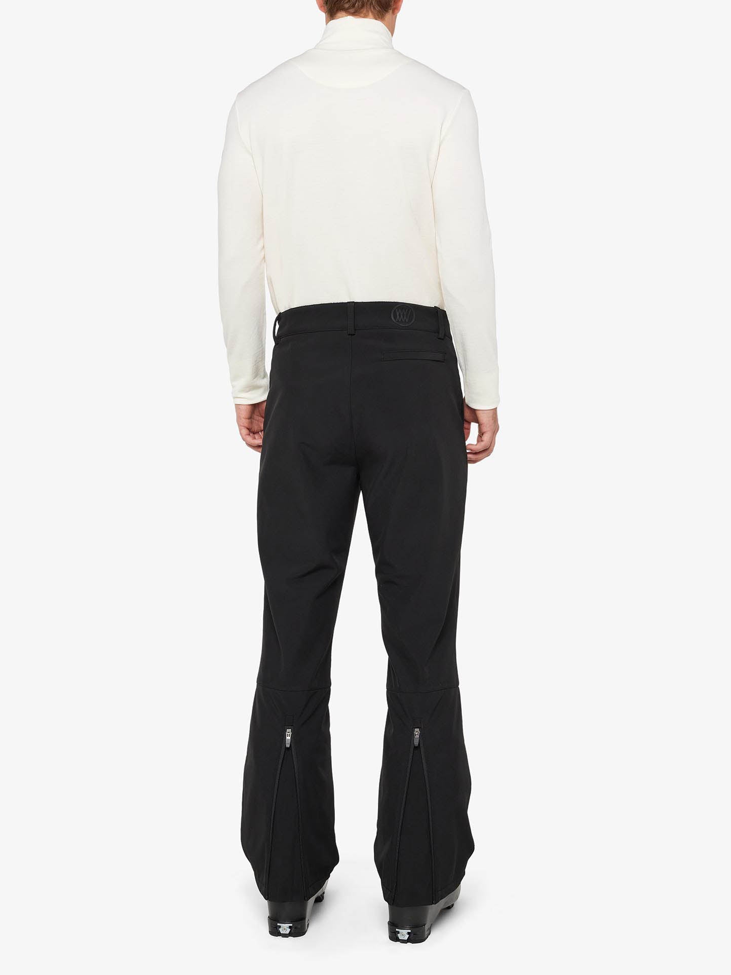 Rear view of a person in a white long-sleeve top and the Tyin Ski Pant Men by We Norwegians, a pair of black high-waisted, slim fit pants with zippers on the back of the legs. The outfit, completed with black boots, is stylishly set against a plain white background.