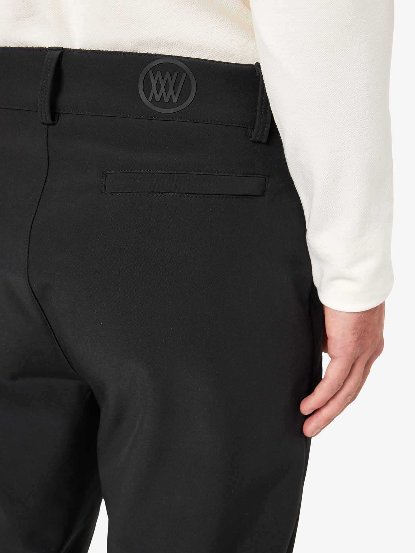 A close-up image of a person wearing the Tyin Ski Pant Men by We Norwegians: sleek, black, slim-fit pants paired with a white top. The pants feature an embroidered "XXV" logo in black near the back belt loop. The person's hand is seen resting on their right hip.