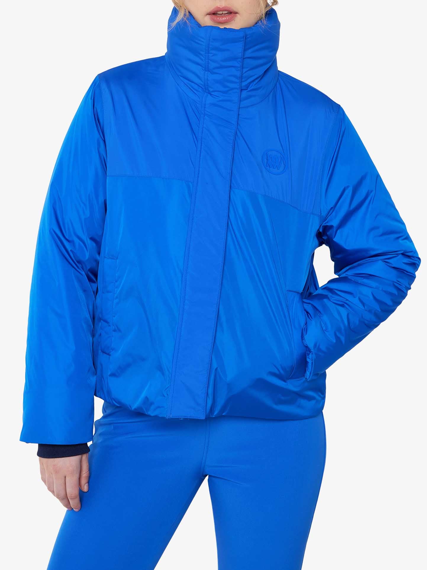 A person wearing a We Norwegians Filefjell Ski Jacket Women in bright blue, with water-repellent fabric and a high collar, stands against a white background. The jacket, featuring wool padding and a logo on the left side of the chest, partially obscures the person's face. They are also wearing matching blue leggings to complete the look.