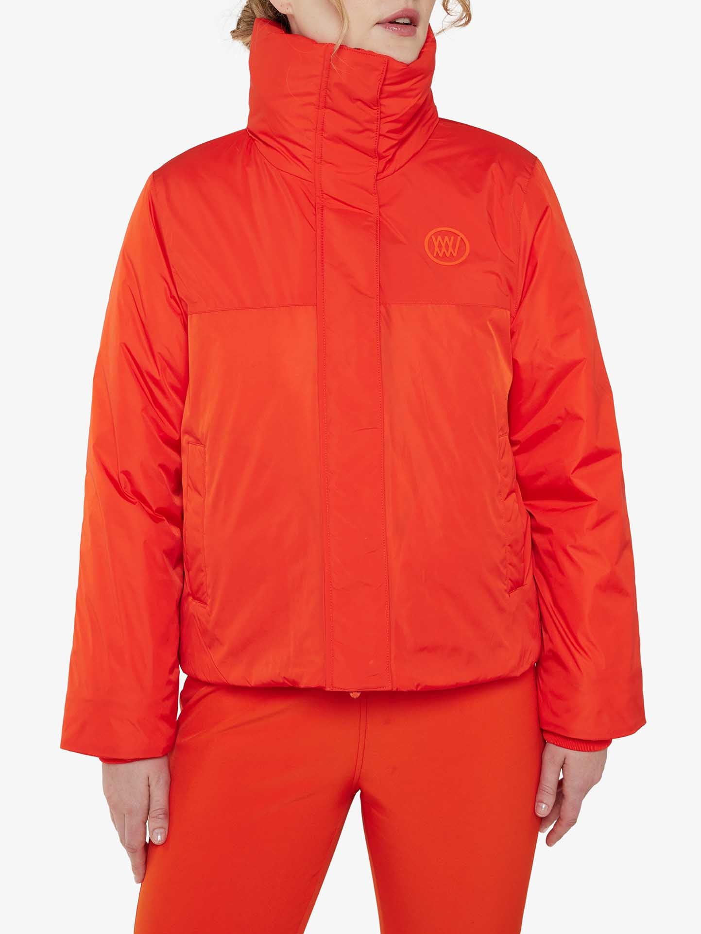 A person is wearing a bright orange Filefjell Ski Jacket Women by We Norwegians, which is water-repellent and features a high collar along with a subtle logo on the upper left side of the chest. They have coordinated this with matching orange pants, and their hair is partially visible against the plain white background.