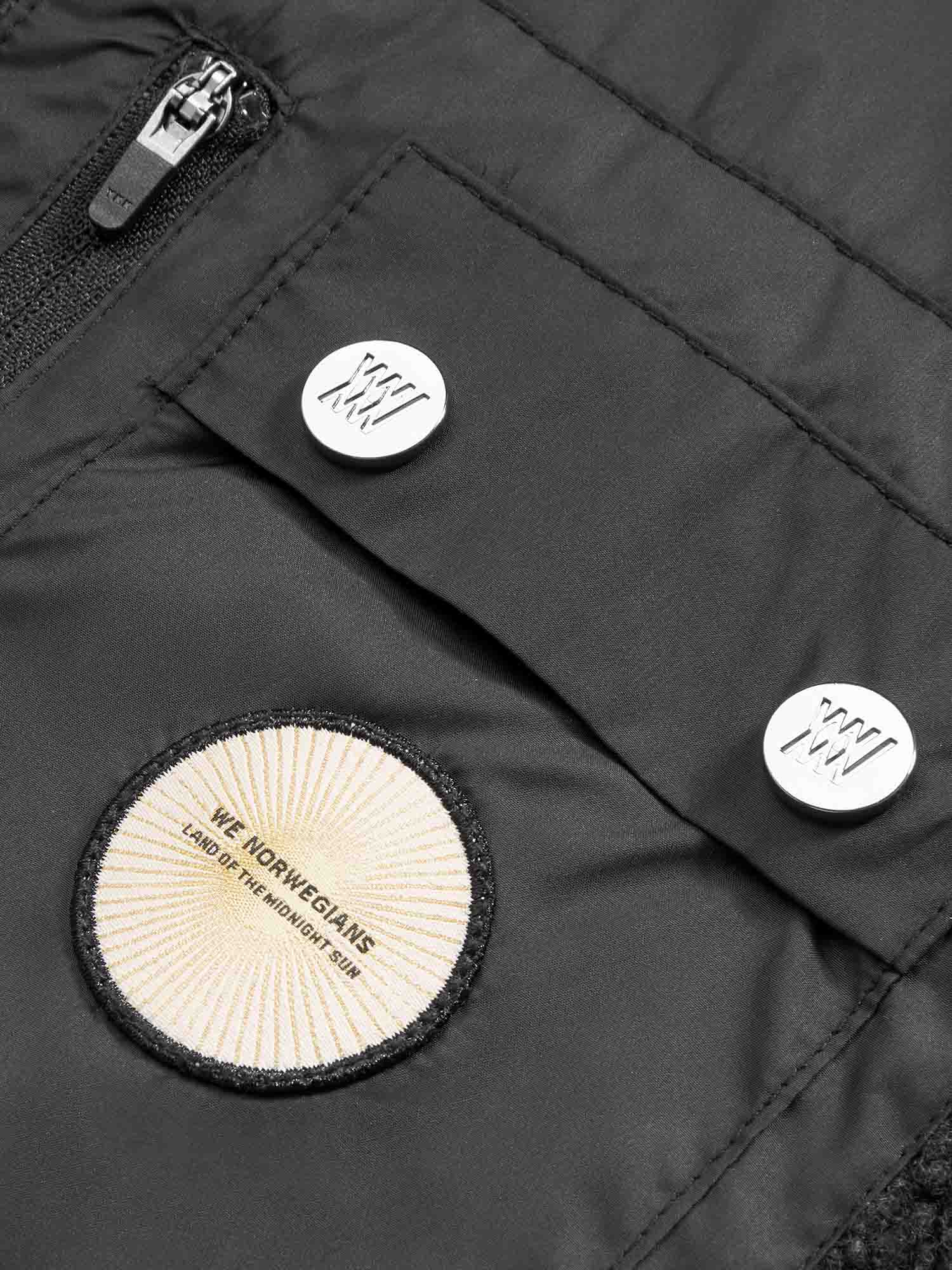 A close-up of a black Alta Jacket Men from We Norwegians, featuring two white buttons with wave-like designs and a circular beige patch. The patch has text at the center that reads "We Norwegians" above smaller text that says "Land of the Northern Light." Ideal for any outdoor activity.