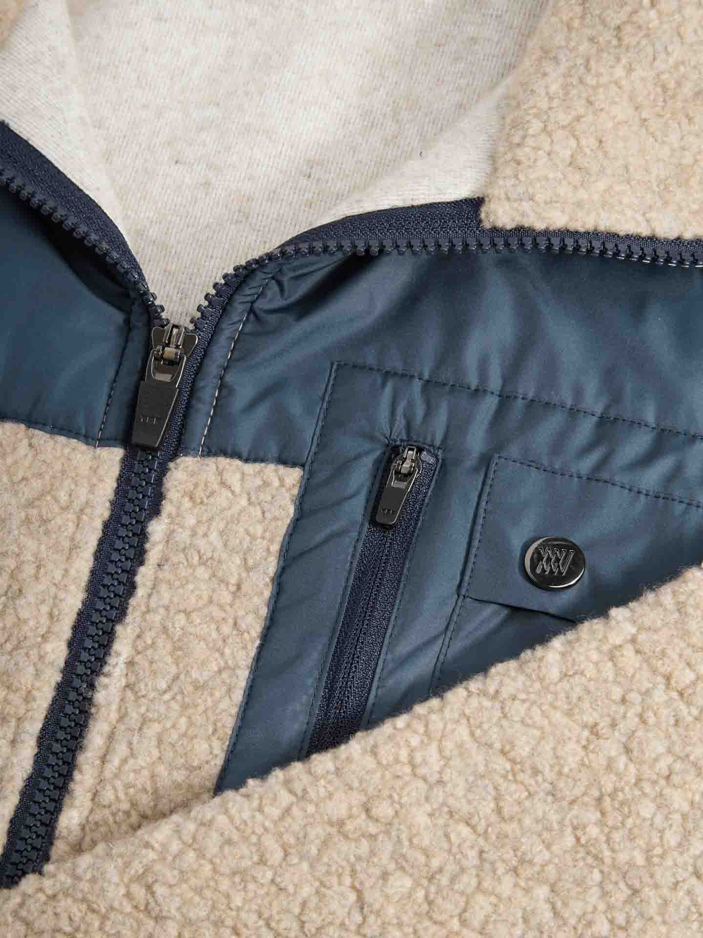 A close-up image of the We Norwegians Alta Jacket Men, showcasing its beige fleece with dark blue nylon trim and zipper. This jacket includes a shiny dark blue pocket with a smaller zipper and a logo button, making it perfect for any outdoor activity. The inner lining is crafted from light-colored fabric.